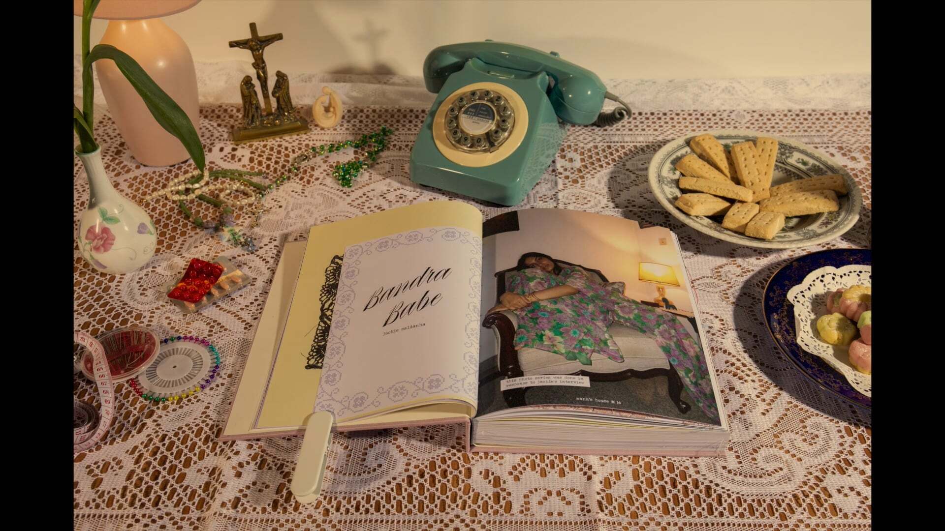 Open pink book with woman sat in sari and text, placed upon an elegant dining table cover alongside a rotary phone, biscuits and trinkets, dim warm light.