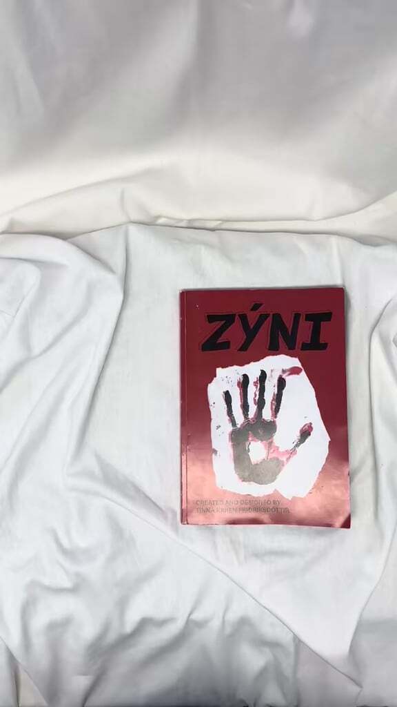 Red book with "ZYNI" title and dark handprint on white background, placed on wrinkled white bedding.