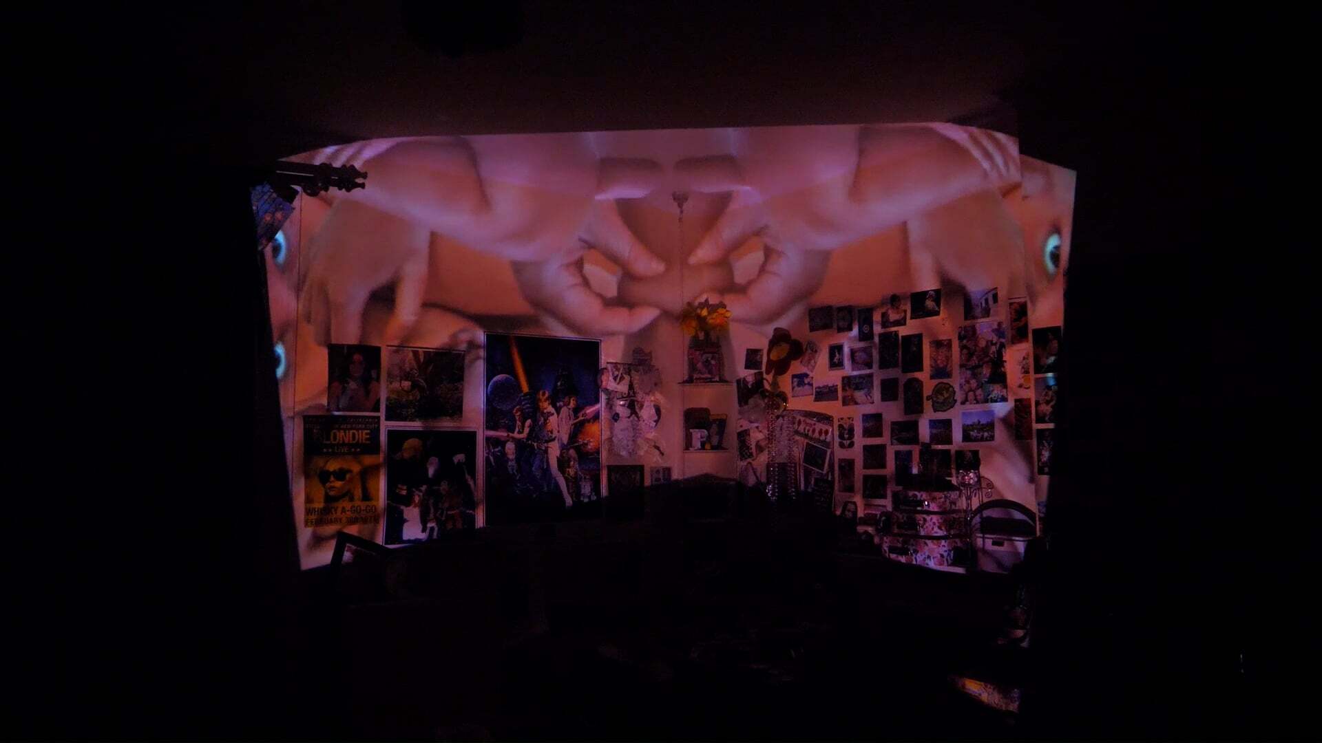 Room interior with pink and purple projection, walls covered in photographs and posters, dark ceiling with decorative elements.