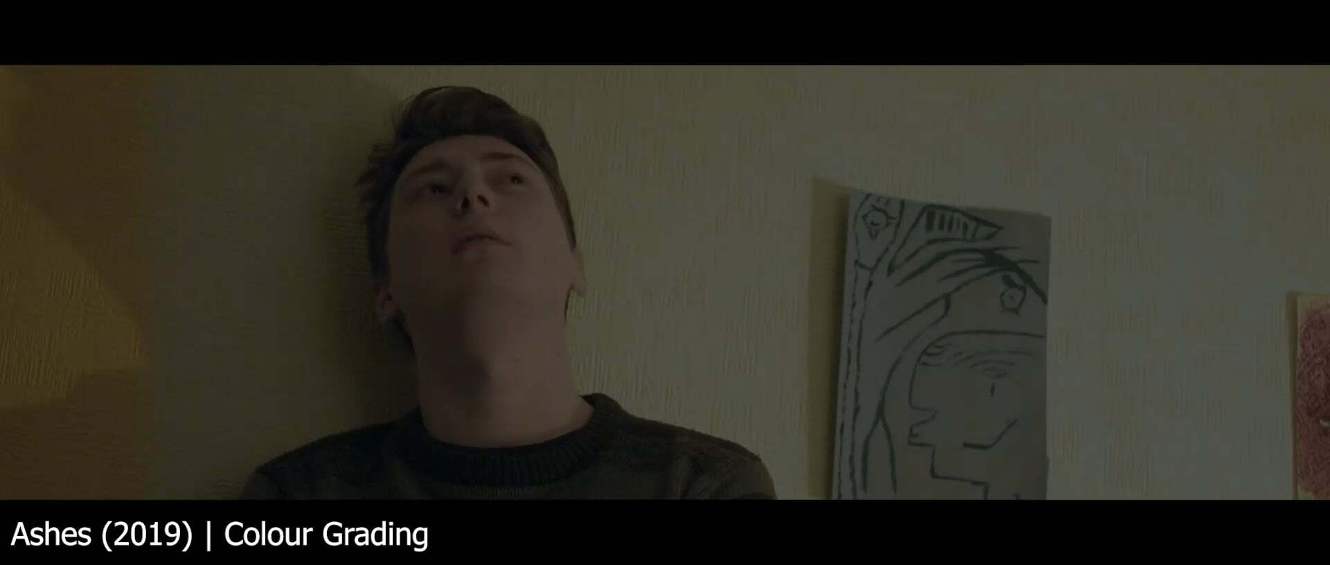 Man in dark shirt looking upwards in dimly lit room with framed artwork on wall behind him.