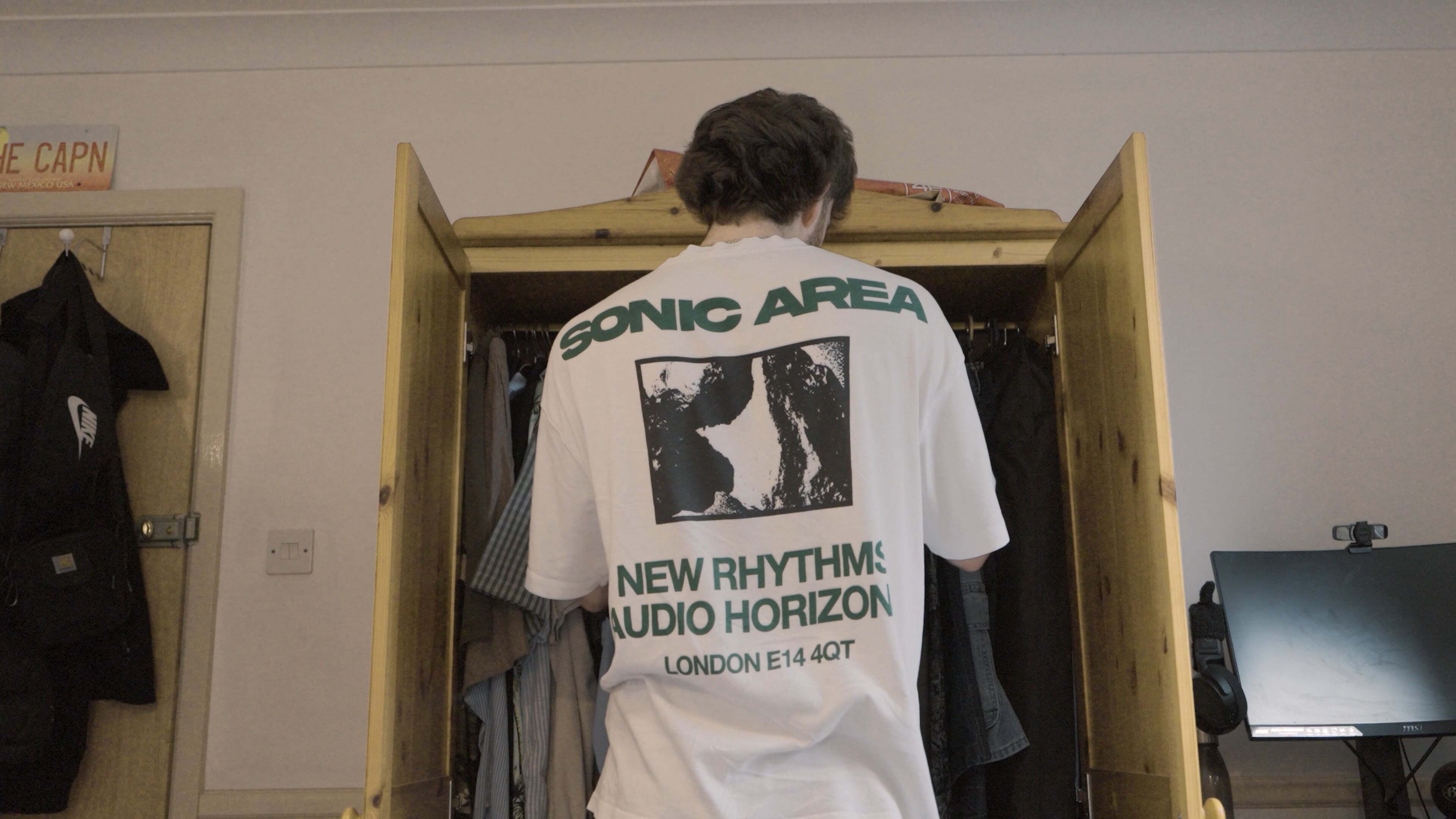 Person in white t-shirt with "SONIC AREA NEW RHYTHM AUDIO HORIZON LONDON" text and black/white image, facing wooden wardrobe.