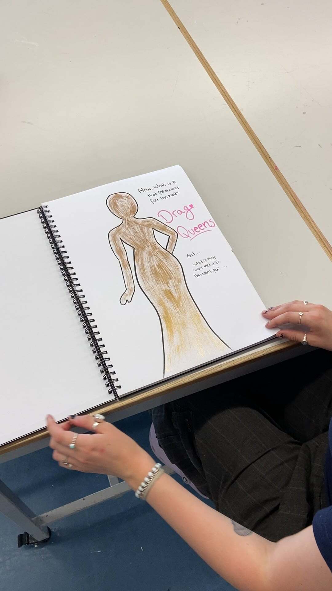 Hands holding open spiral notebook with brown and yellow coloured drawing of female figure, pink text "Days Queen" visible on white page.