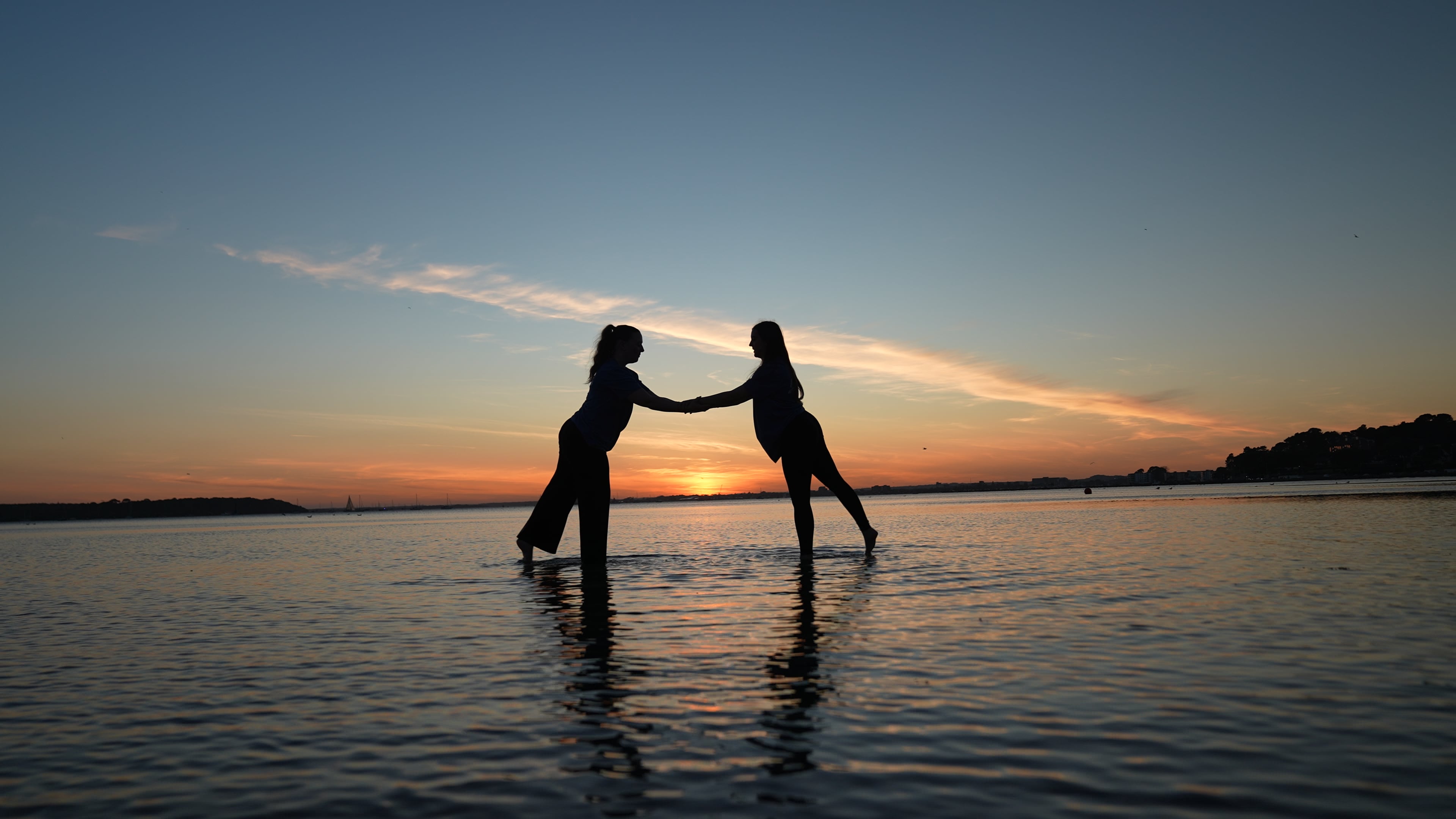 Two dancers perform on the surface of a body of water at sunset. One dancer bends forward with one leg raised, while the other stands on one leg, extending their arms, against a backdrop of orange and purple hues in the sky.
