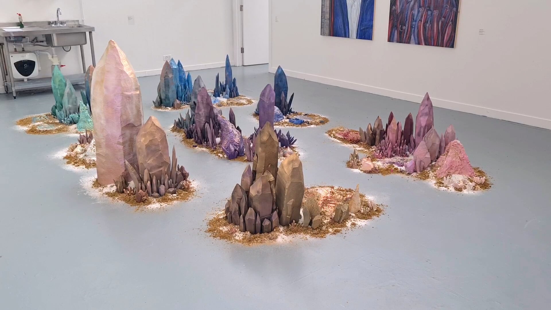 An installation of large, colourful crystal formations placed on the floor of an art gallery. The crystals vary in height and colour, including shades of pink, purple, blue, and green, arranged amidst a bed of white material resembling salt.