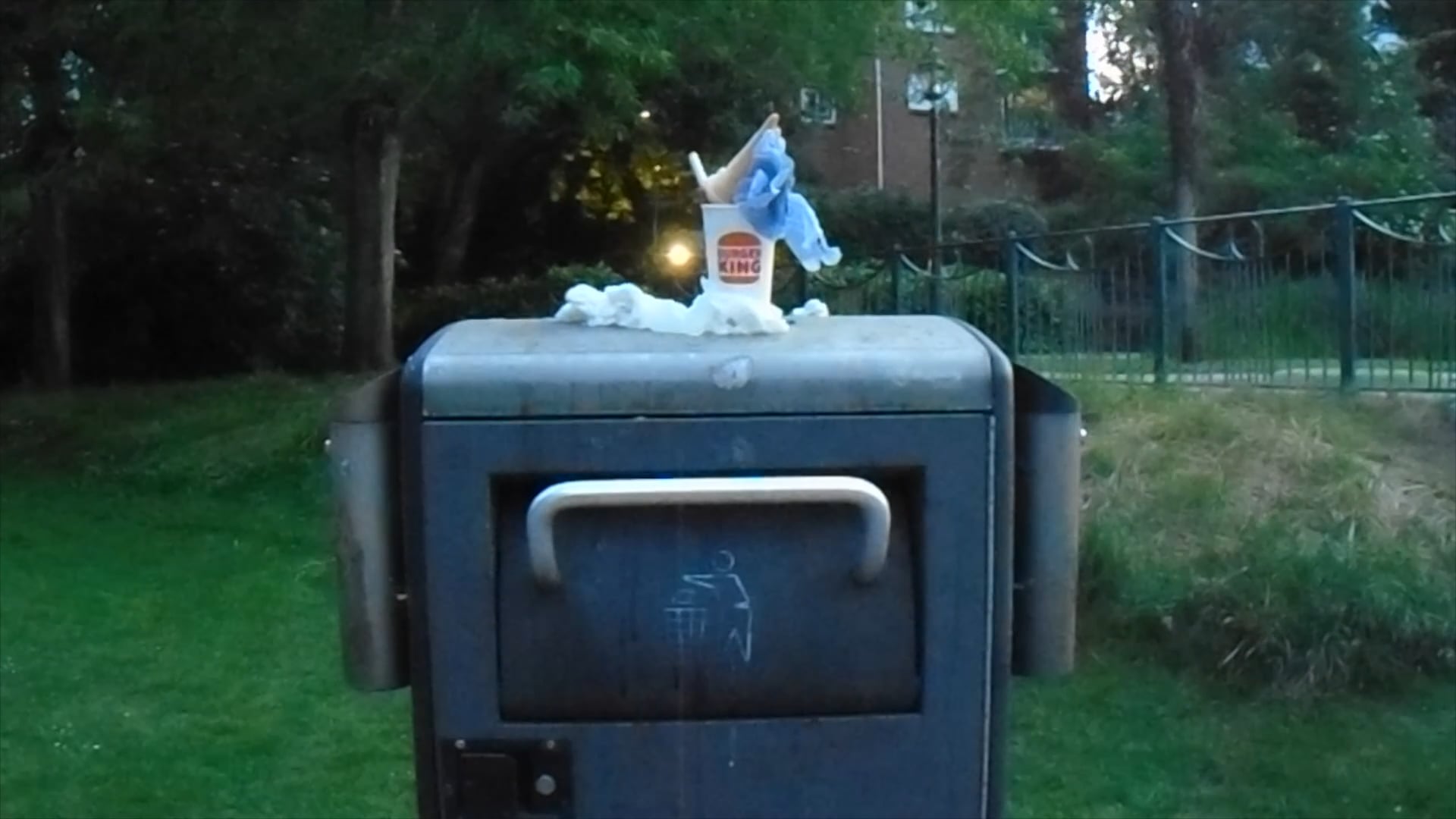 A grey metal bin is topped with a discarded fast-food container and some crumpled paper. In the background, greenery and park fencing are visible under the evening light.