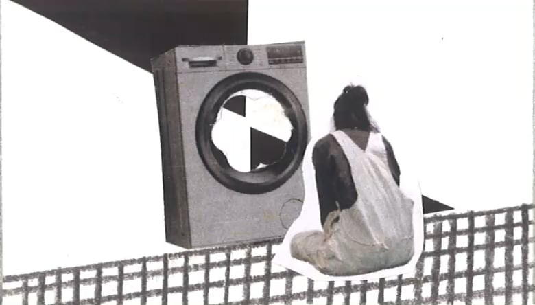 A person in light overalls sits facing a large washing machine sculpture, with a black-and-white geometric backdrop and grid fence behind.