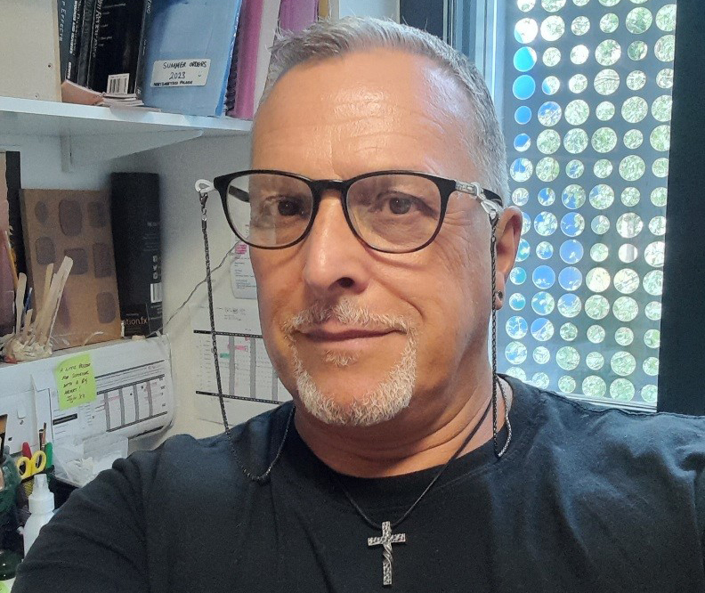 Man with grey hair and beard wearing black-rimmed glasses and black shirt with cross necklace in office with shelves and circular wall decoration.