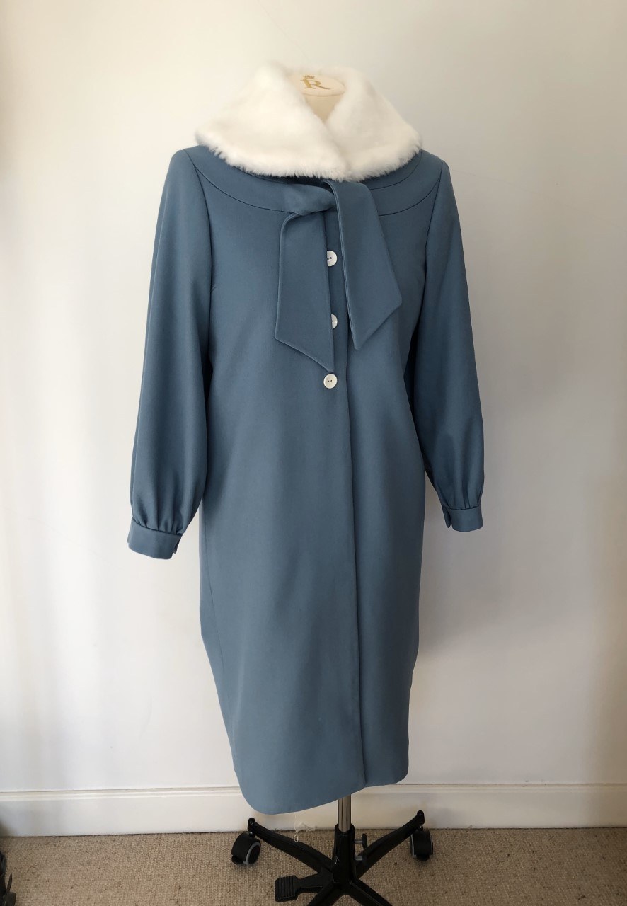 A coat with a fur collar hangs on a mannequin against a plain wall.