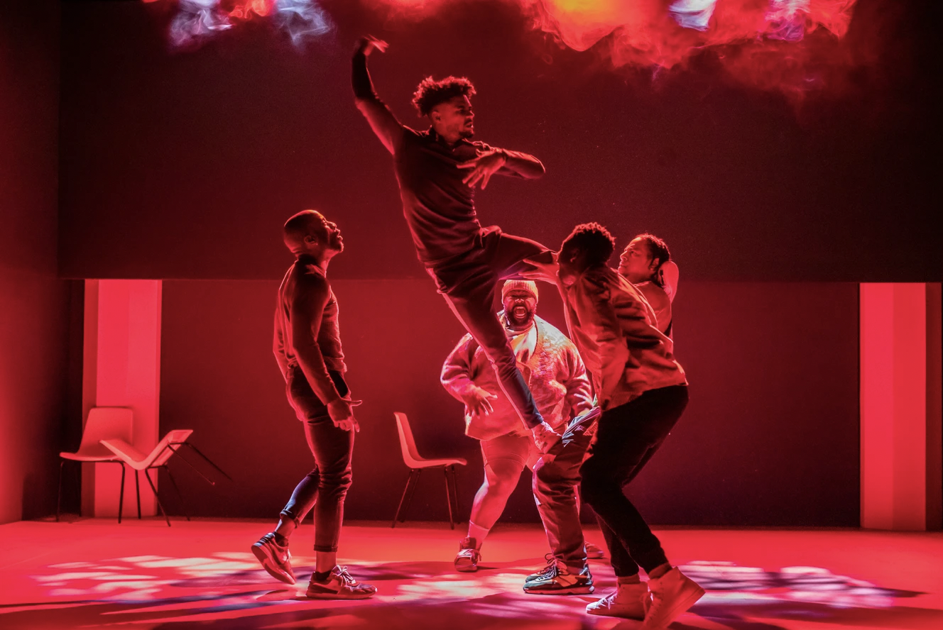 Actors on-stage under red lighting, one in the middle being launched into the air in a pose surrounded by four others.