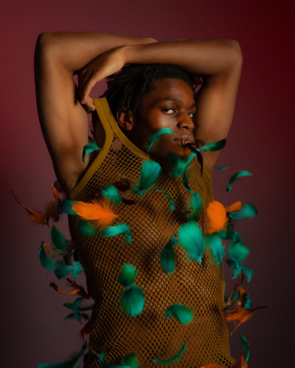 A male model wearing green and orange feathers on a dark red background.