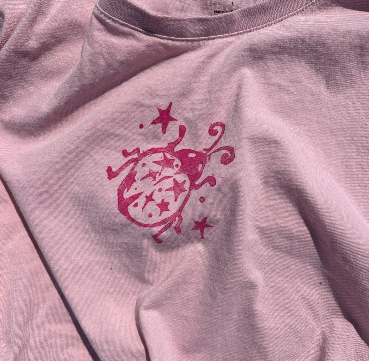 A close-up of a soft pink T-shirt featuring a printed design of a stylised beetle adorned with stars and swirls in a darker pink shade. The fabric appears slightly wrinkled, and the neckline is a traditional crew style.