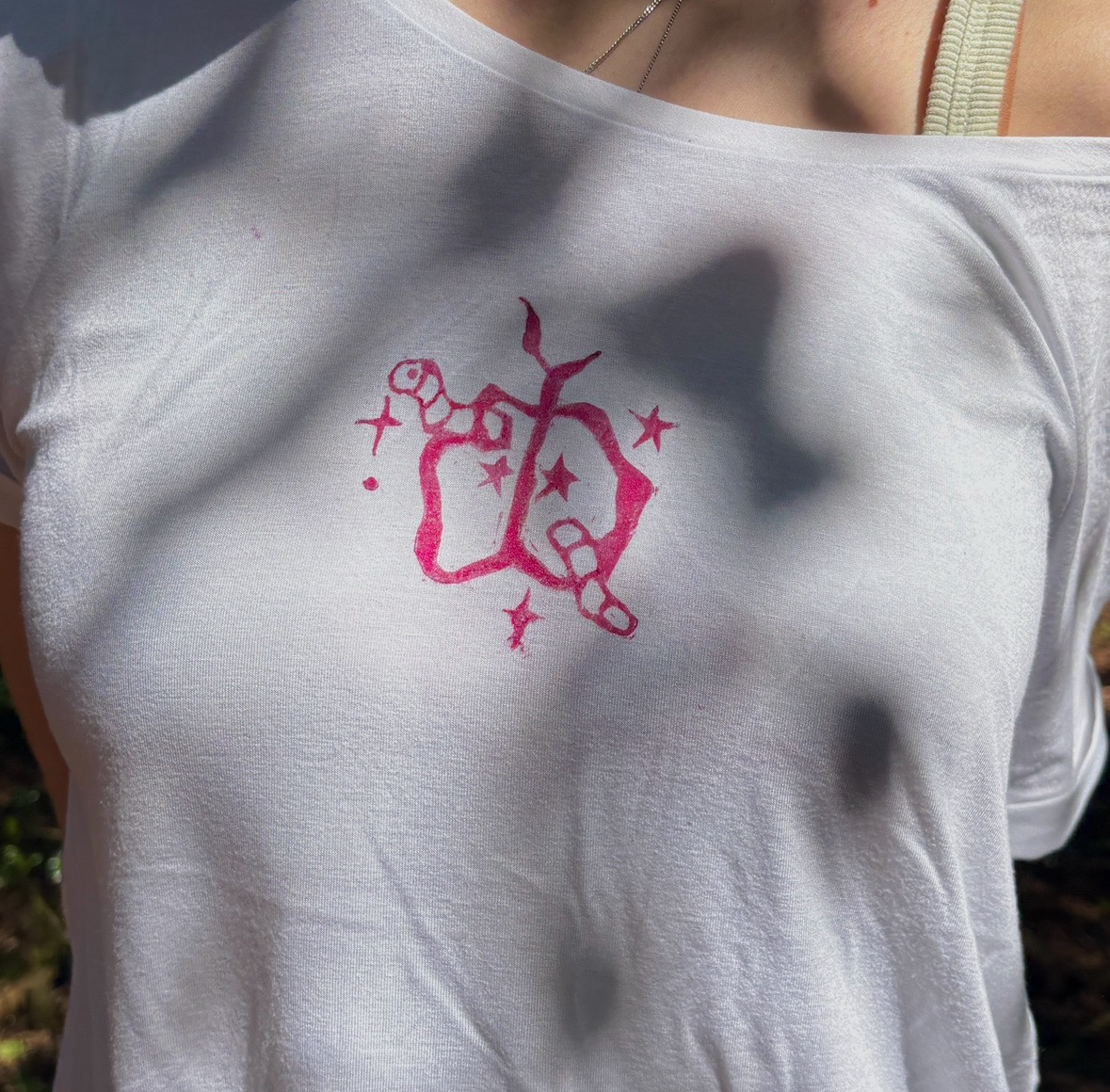 A close-up of a person's upper body wearing a white off-shoulder shirt. The shirt features a pink graphic design in the centre, which includes intertwined shapes and stars.