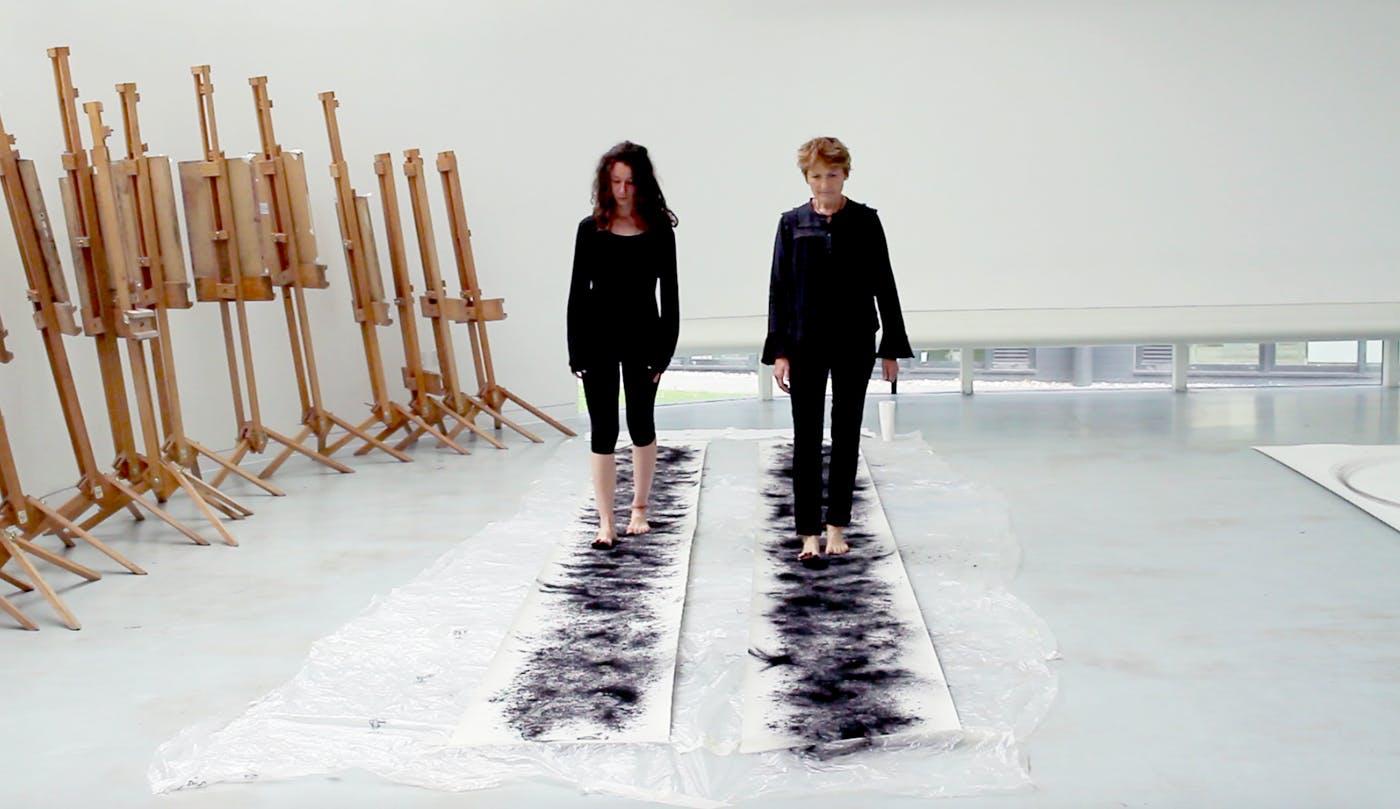 Two women walk on top of two long pieces of paper with heavy charcoal marks on them.