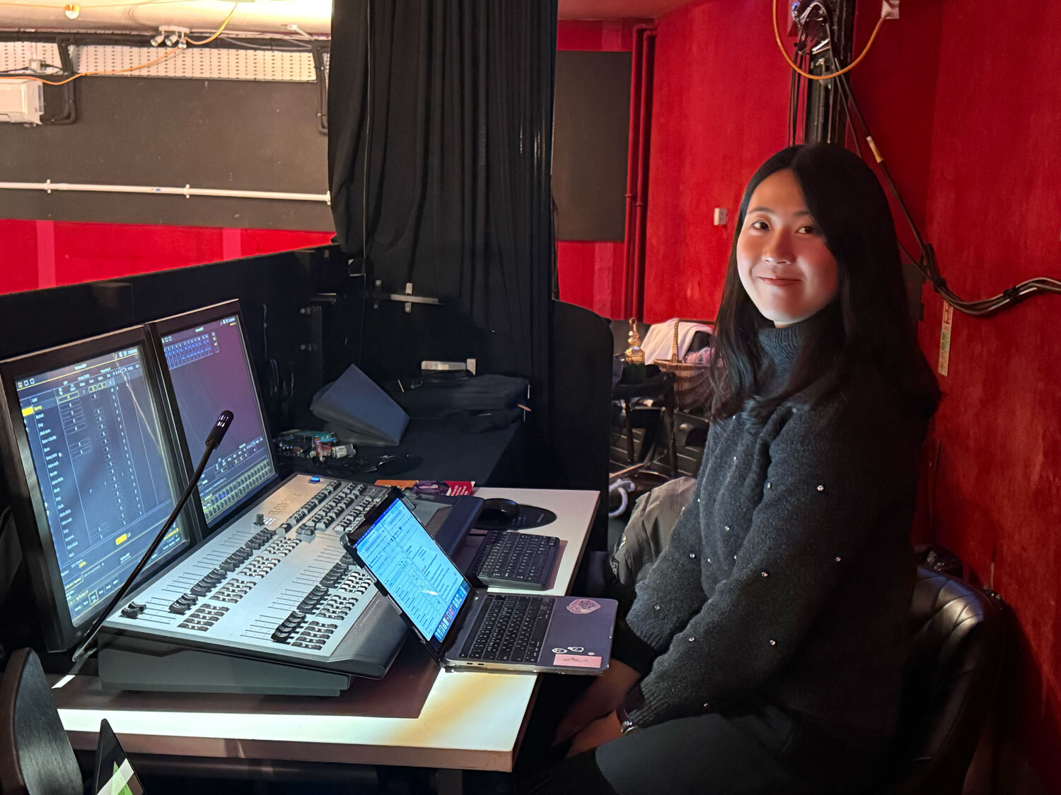 AUB student seated at radio broadcasting desk with mixing console, computer monitors, and microphone in red-walled studio.