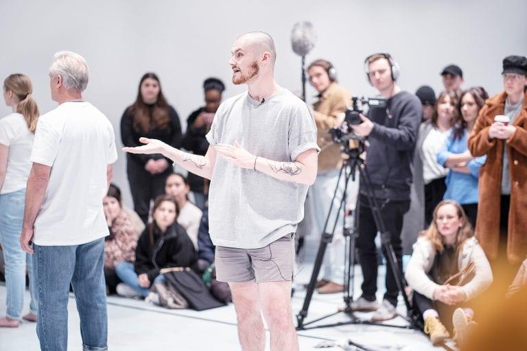 A group of people gather in an open studio or gallery space. One person is shown standing in front of a camera man.