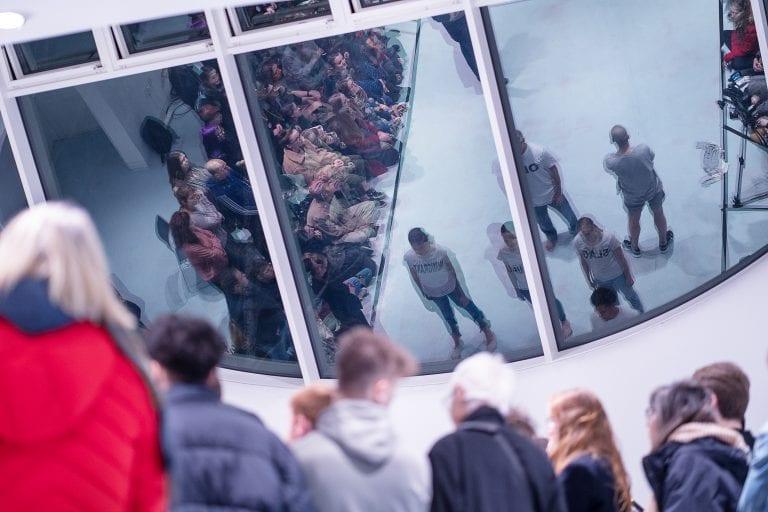 A group of people gathers with a series of windows and their reflections above them.