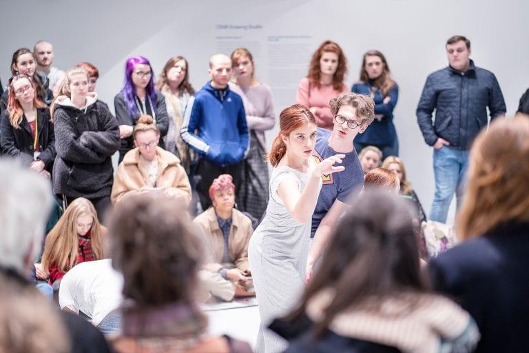 A crowd gathers around and watches a pair of performers in a gallery space.