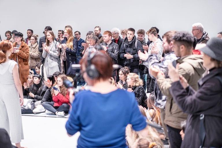 A crowd of people gather around a performer taking photos and videos on cameras and phones.