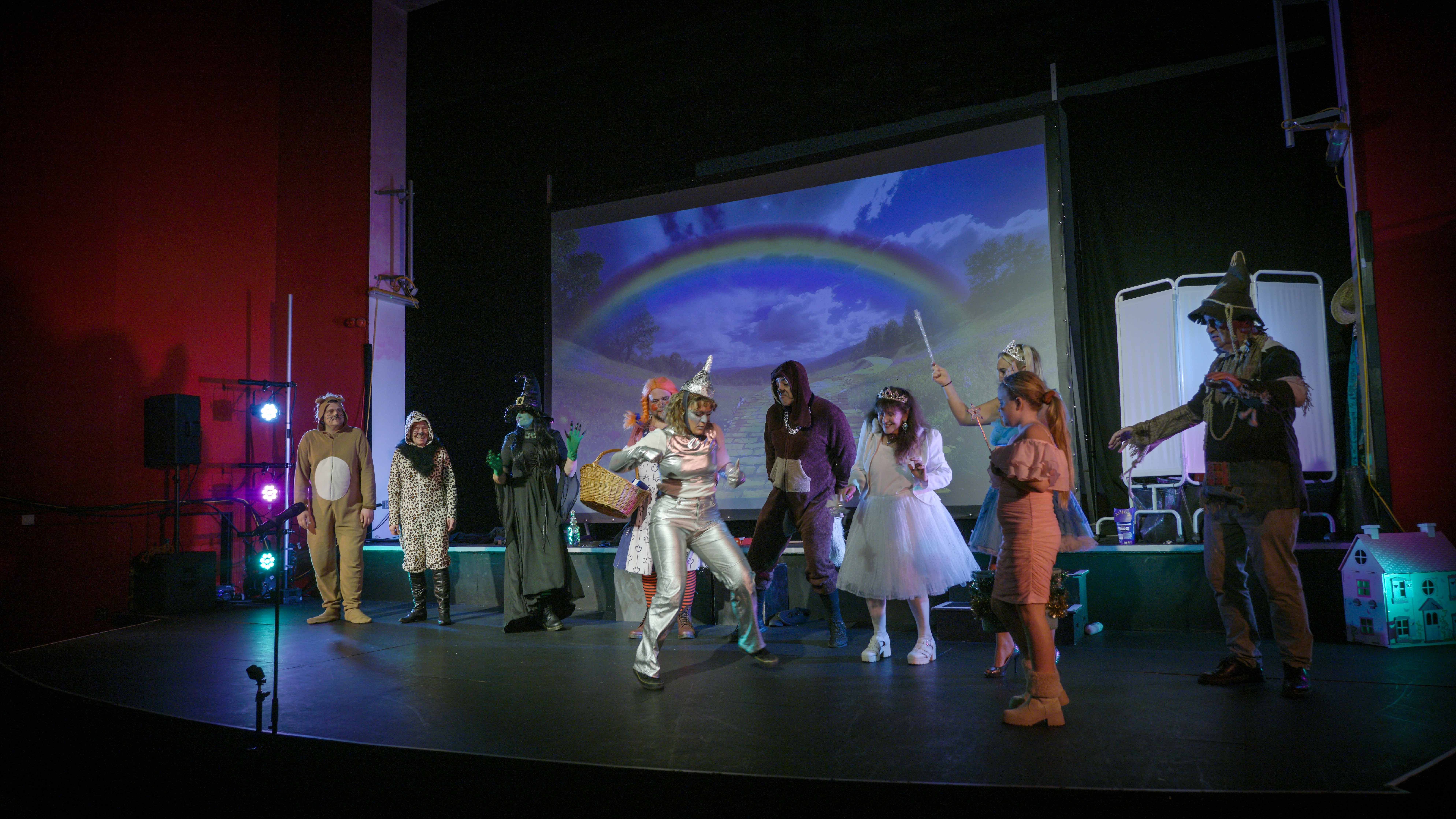 Theatre stage with costumed performers, blue projected backdrop showing rainbow, coloured stage lighting, and audience silhouettes visible.