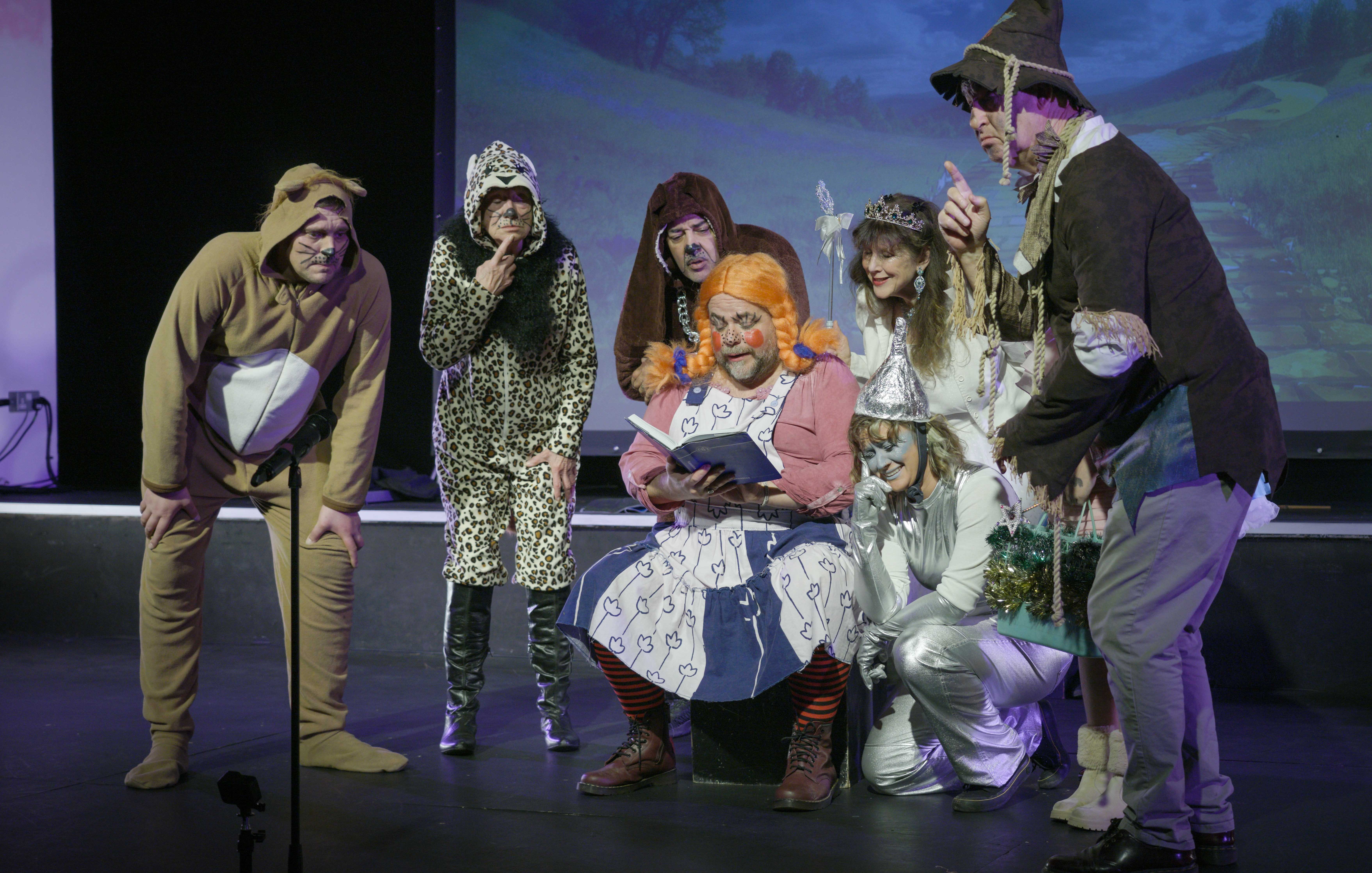 Theatre performers in costume on stage: brown monk robes, leopard print coat, orange jumper, witch hat, white dress, dark suit against blue backdrop.
