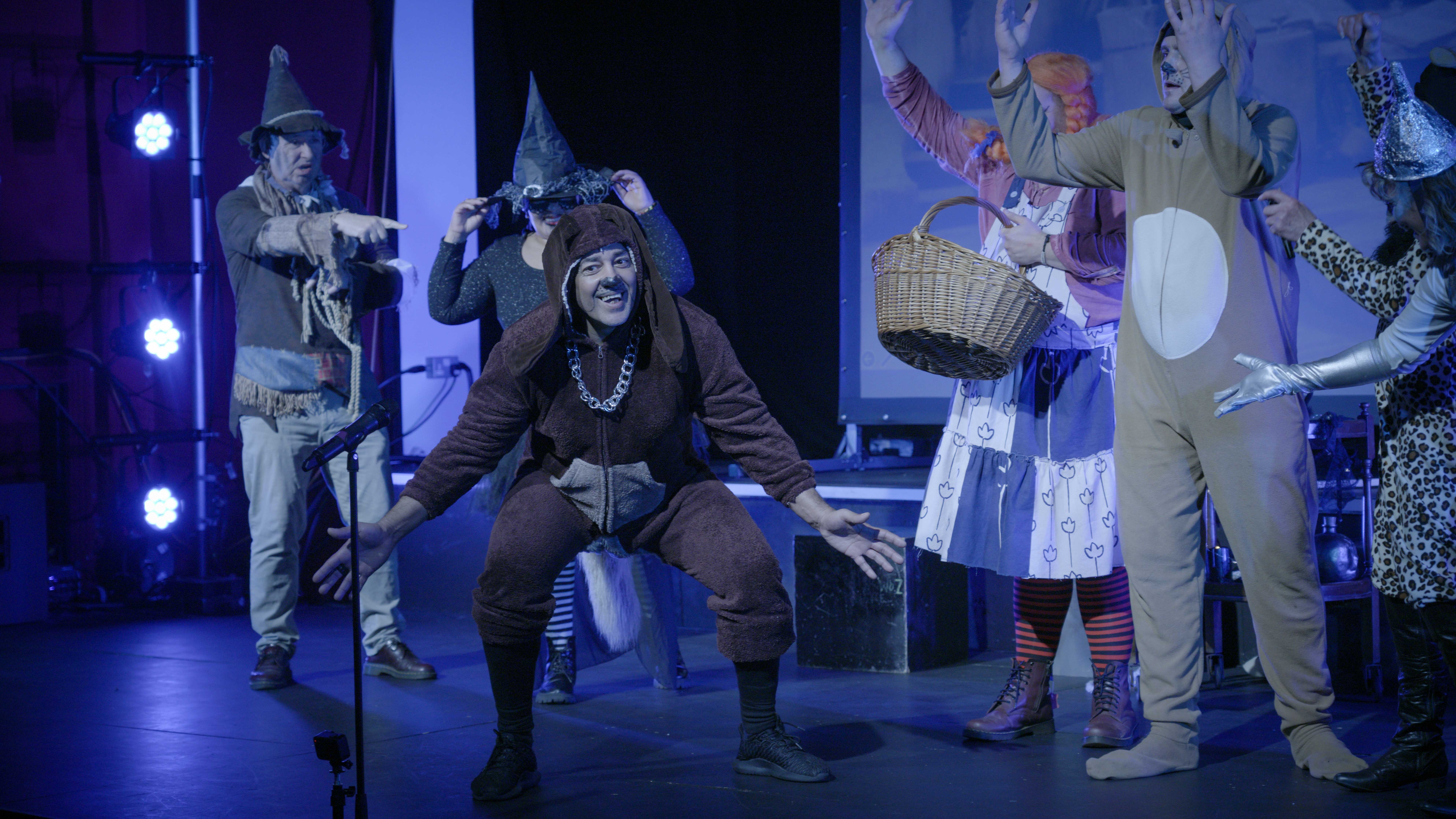 Theatre performers in costume on stage with blue lighting. Central figure in brown outfit crouched down, others in period dress around them.