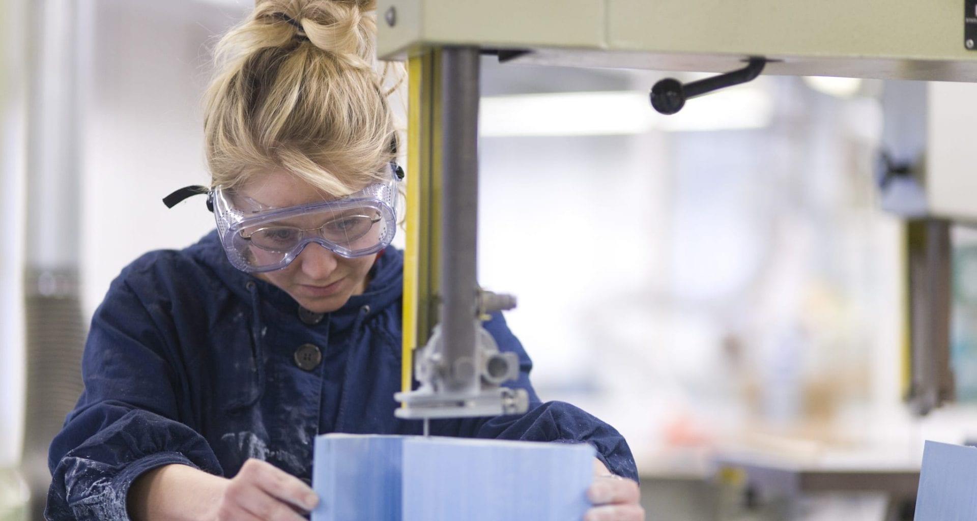 Student wearing safety goggles using a machine in a workshop.