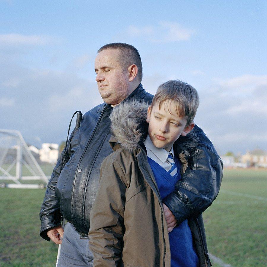 Two people stand on a football pitch. The taller person has their arm around the shorter person.