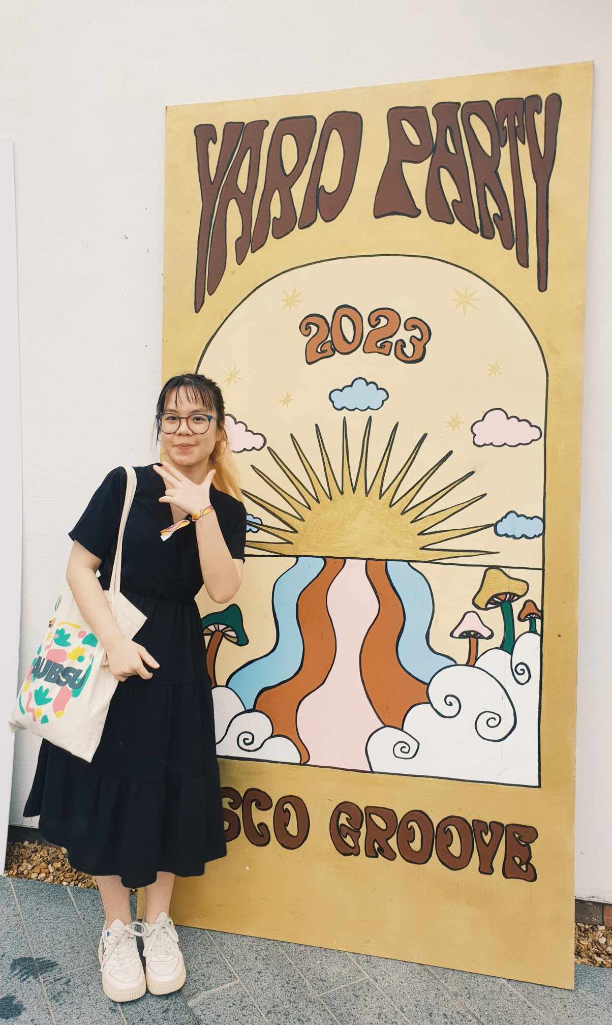 Ngan Nyugen stood besides AUB's brown-themed Yard Party 2023 banner with a framed sun and rainbow illustration.