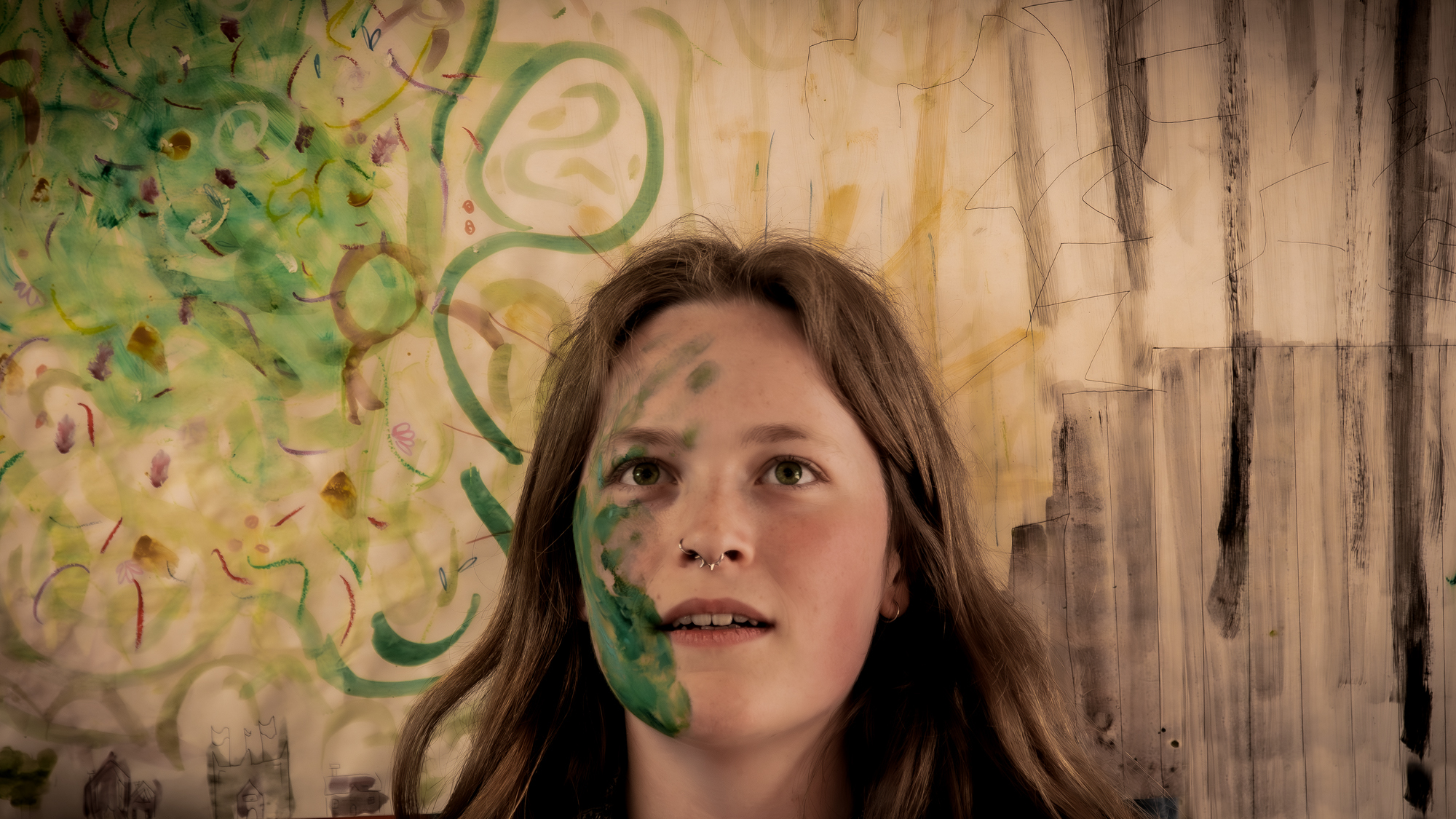 Woman with brown hair looking upwards against wall with green circular graffiti and vertical brown streaks.