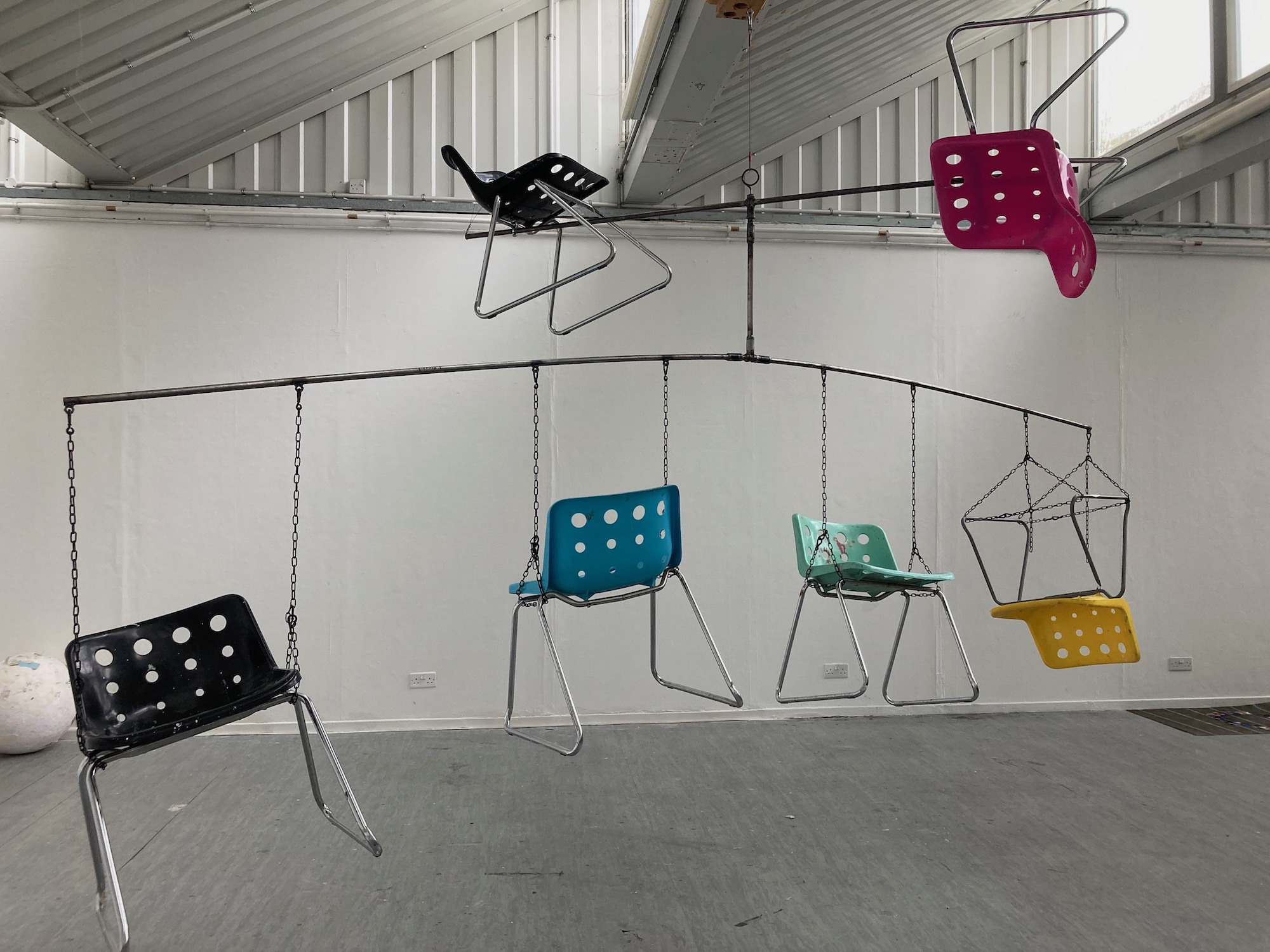 A large mobile with different coloured chairs attached to it, hanging from a gallery ceiling.