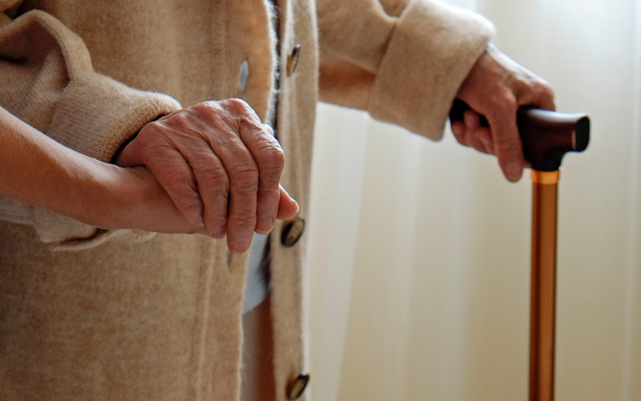 An elderly person walking with a walking stick and the support of another person's hand