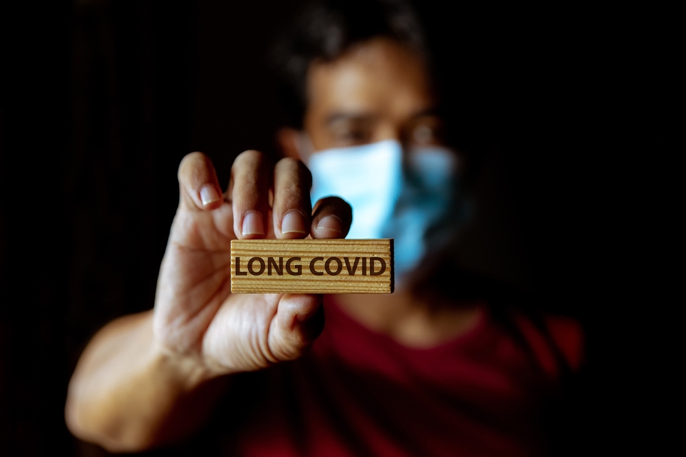 Masked man shows wooden sign with wording "Long Covid" in dark room.