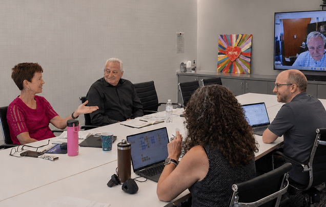 A group of people sitting around a table with a laptop and a TV in the background. Description generated by AI