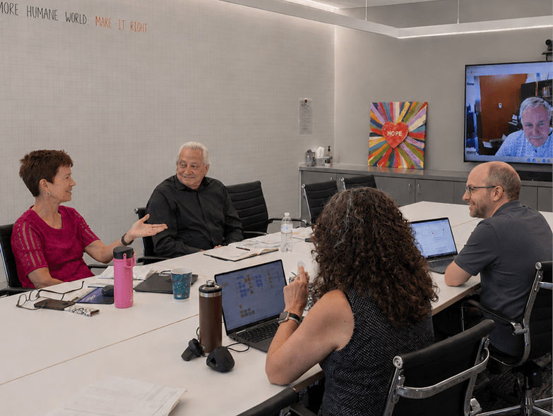 A group of people sitting around a table with a laptop and a TV in the background. Description generated by AI