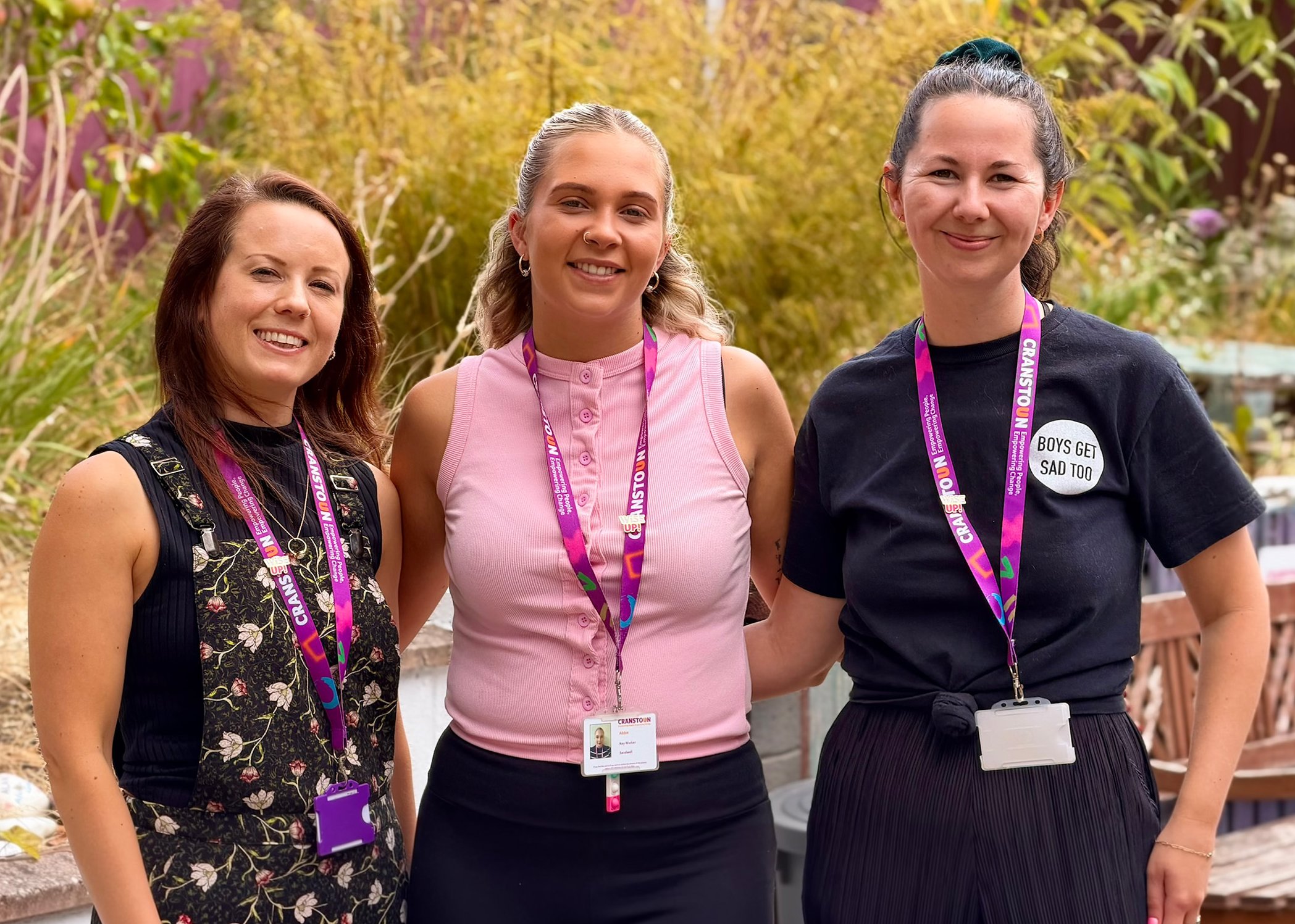 Three women wearing purple Cranstoun lanyards smiling at the camera while standing in a garden.
