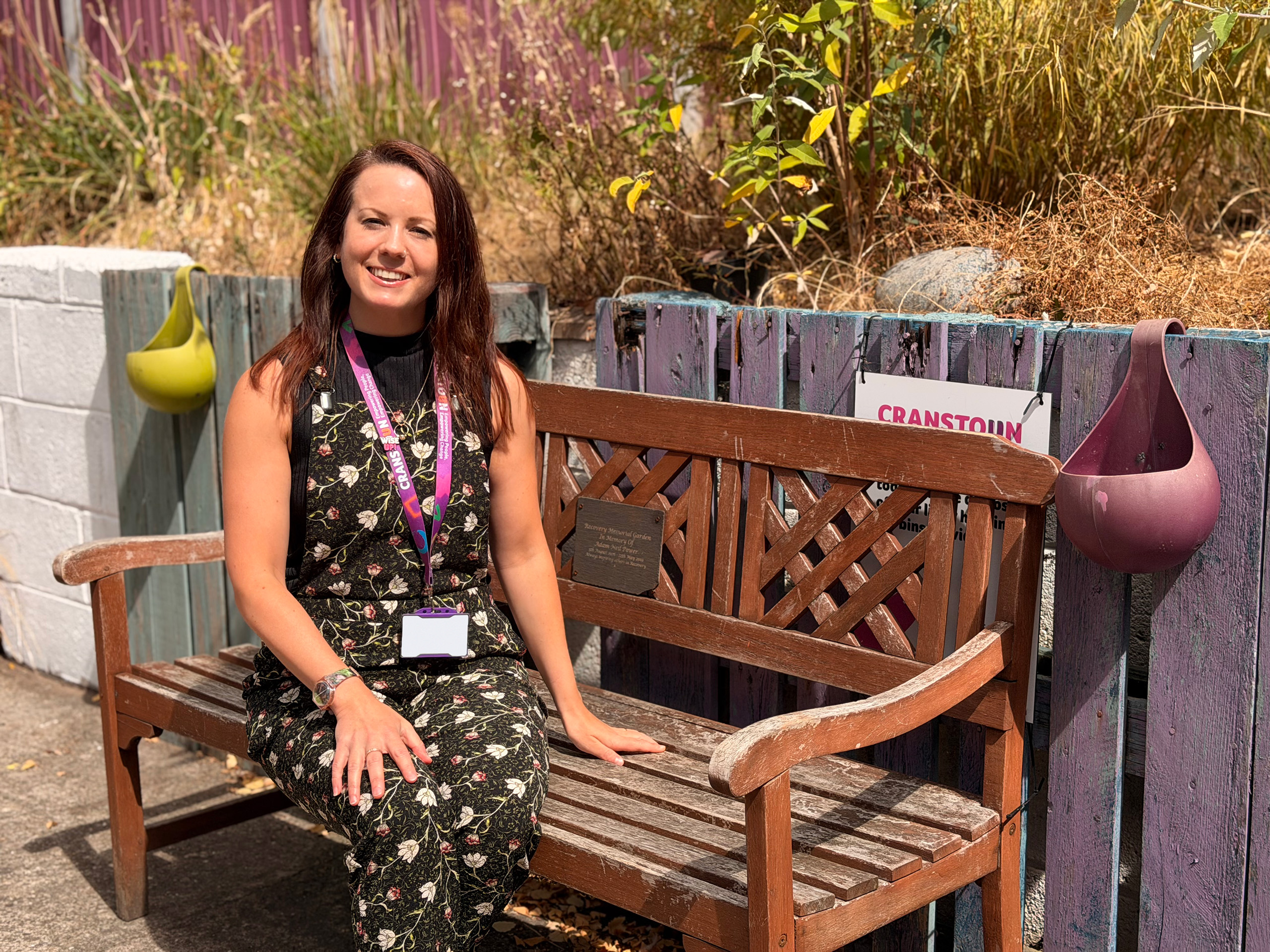 Beth, a woman wearing a black floral jumpsuit and purple Cranstoun lanyard, smiling while sitting on a bench.