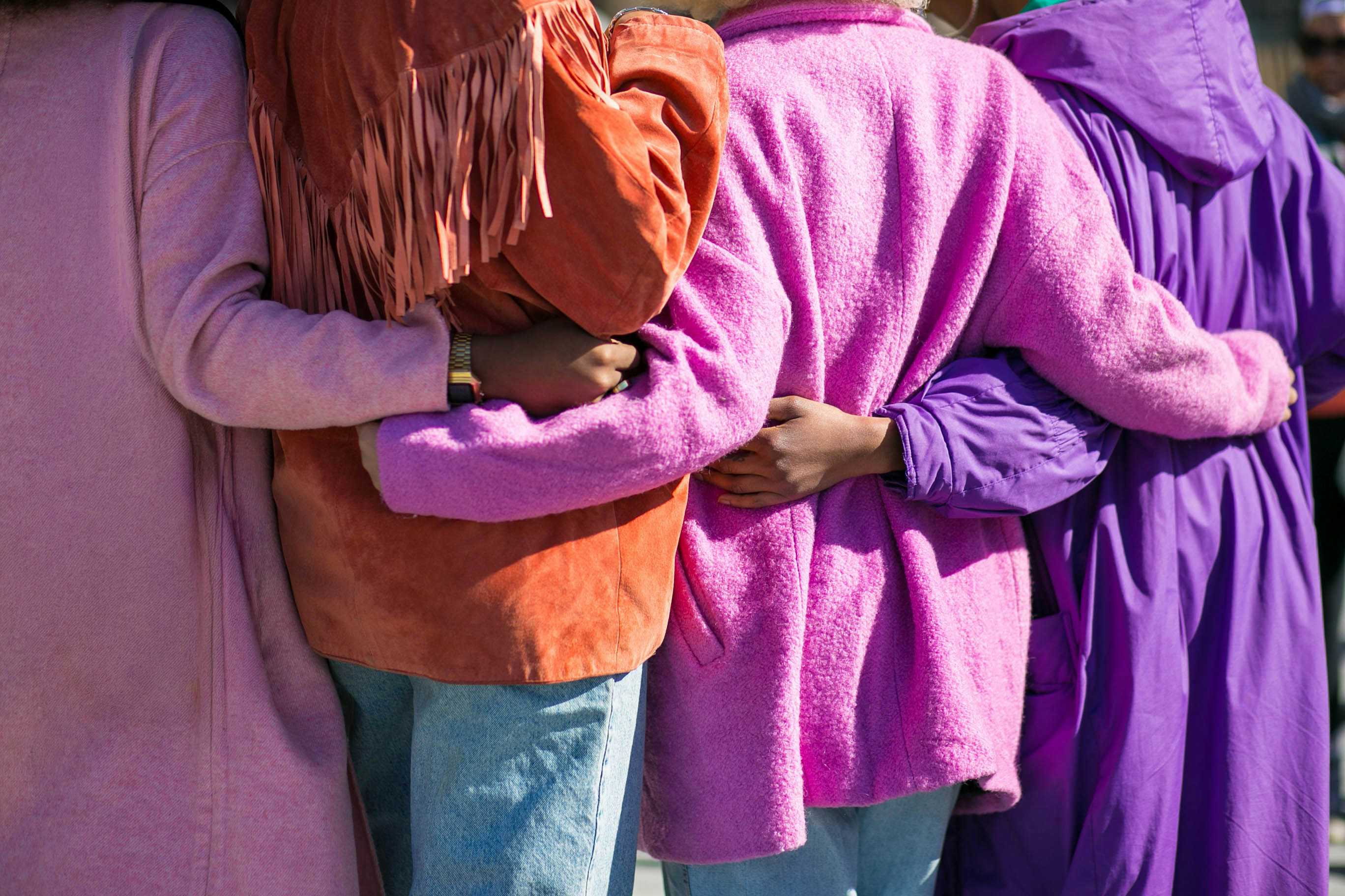 Four women standing side by side with their arms around each other's backs. The image is cropped to show only their torsos and arms, with purple, pink and orange clothing filling most of the frame.