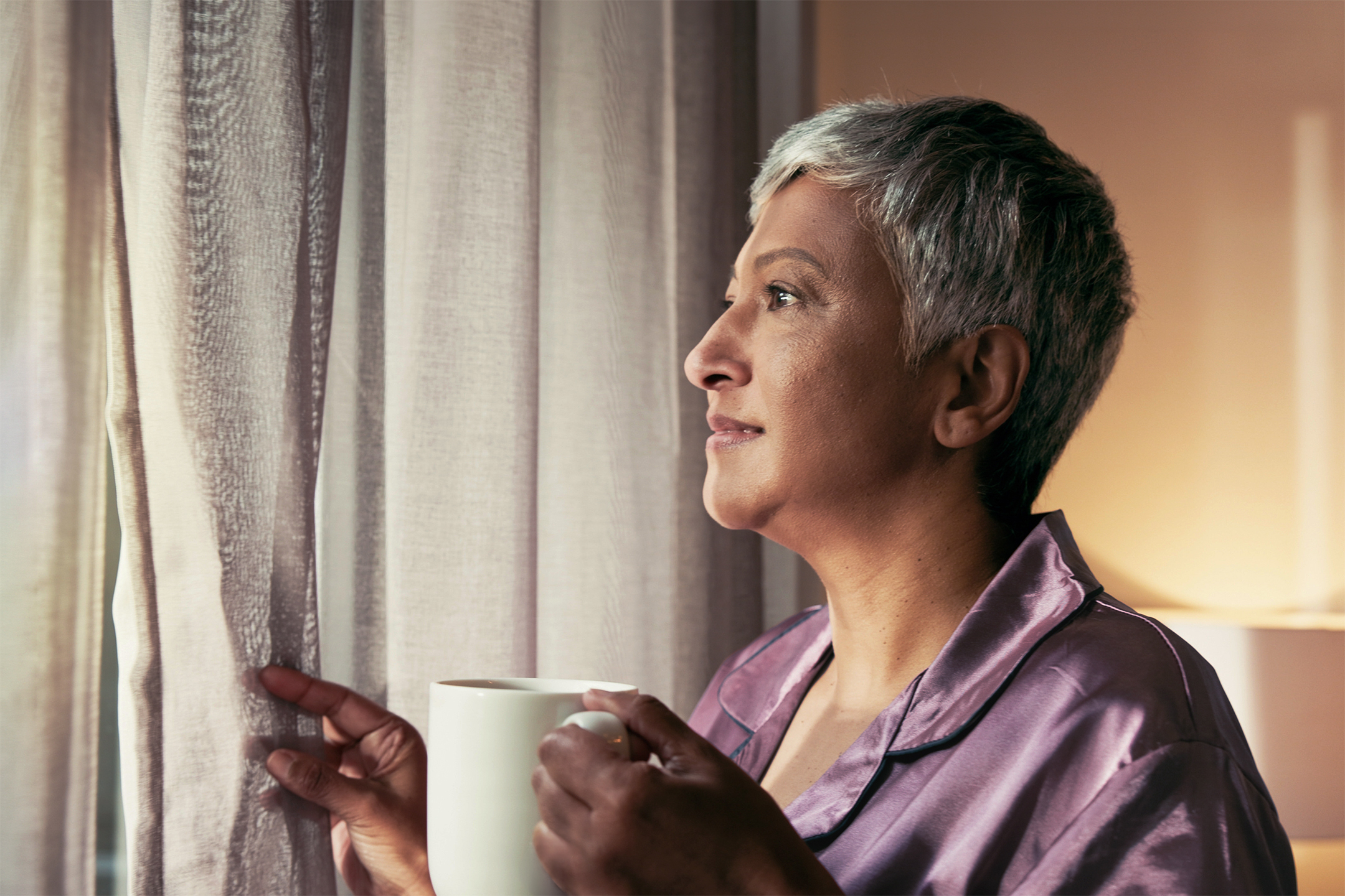A woman in a dressing gown holding a mug of tea, looking out through sheer curtains in a softly lit room.