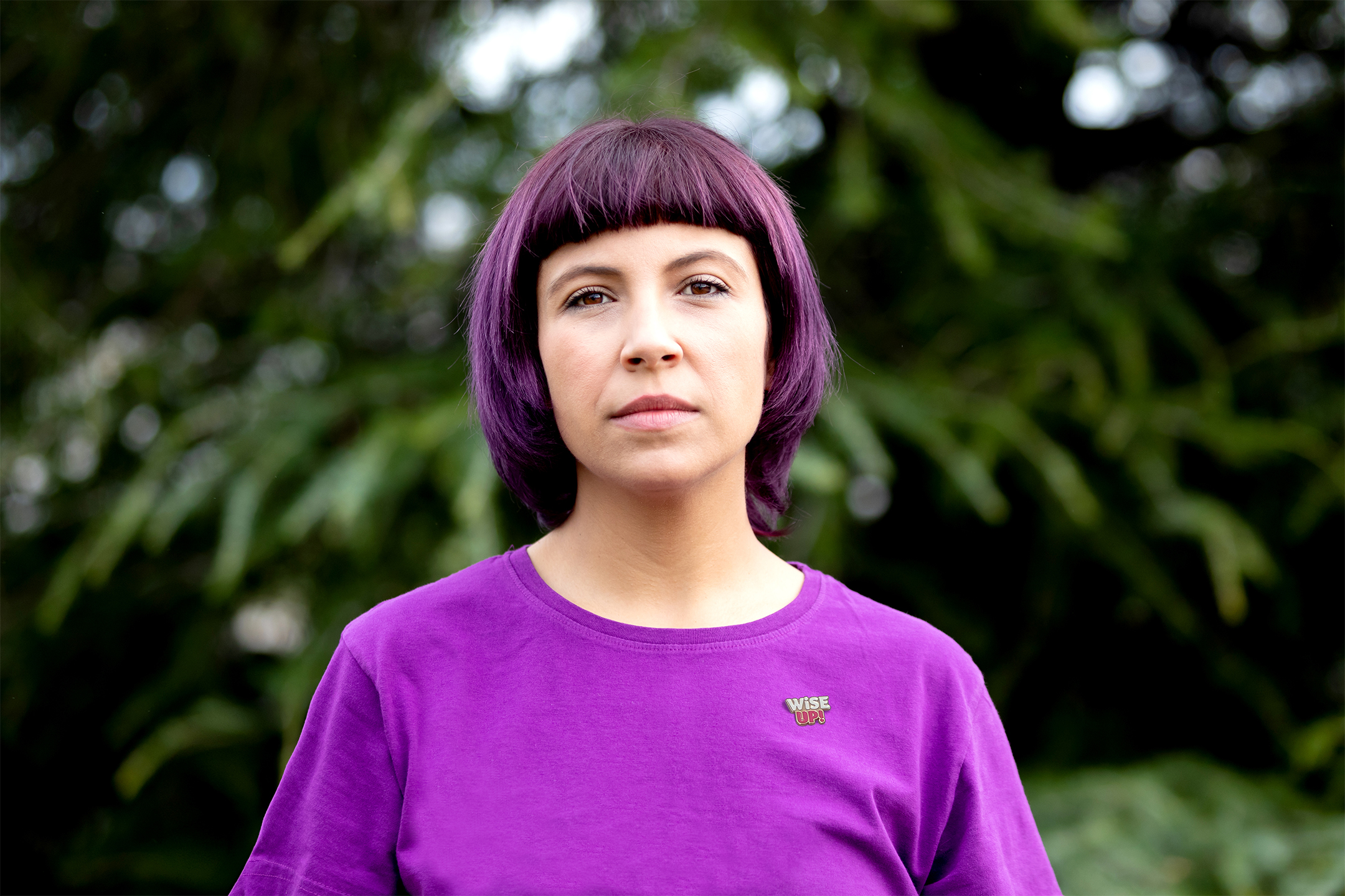 Portrait of a woman with short purple hair wearing a purple T-shirt with a “WISE UP!” pin badge.