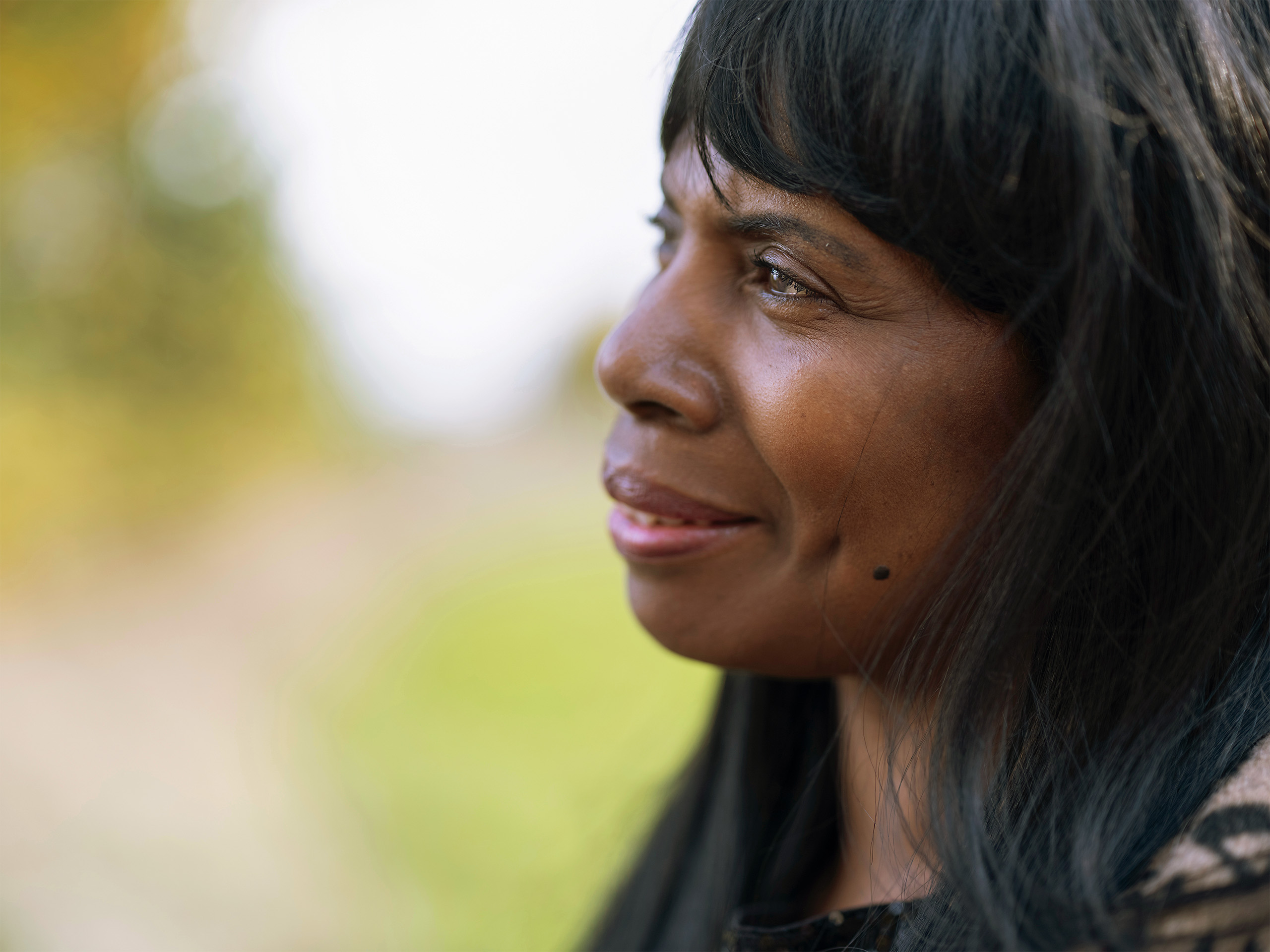 A close-up side profile of a woman outdoors, looking ahead and smiling softly, with a blurred green background. The photo represents a woman accessing support through the WiSE Project.