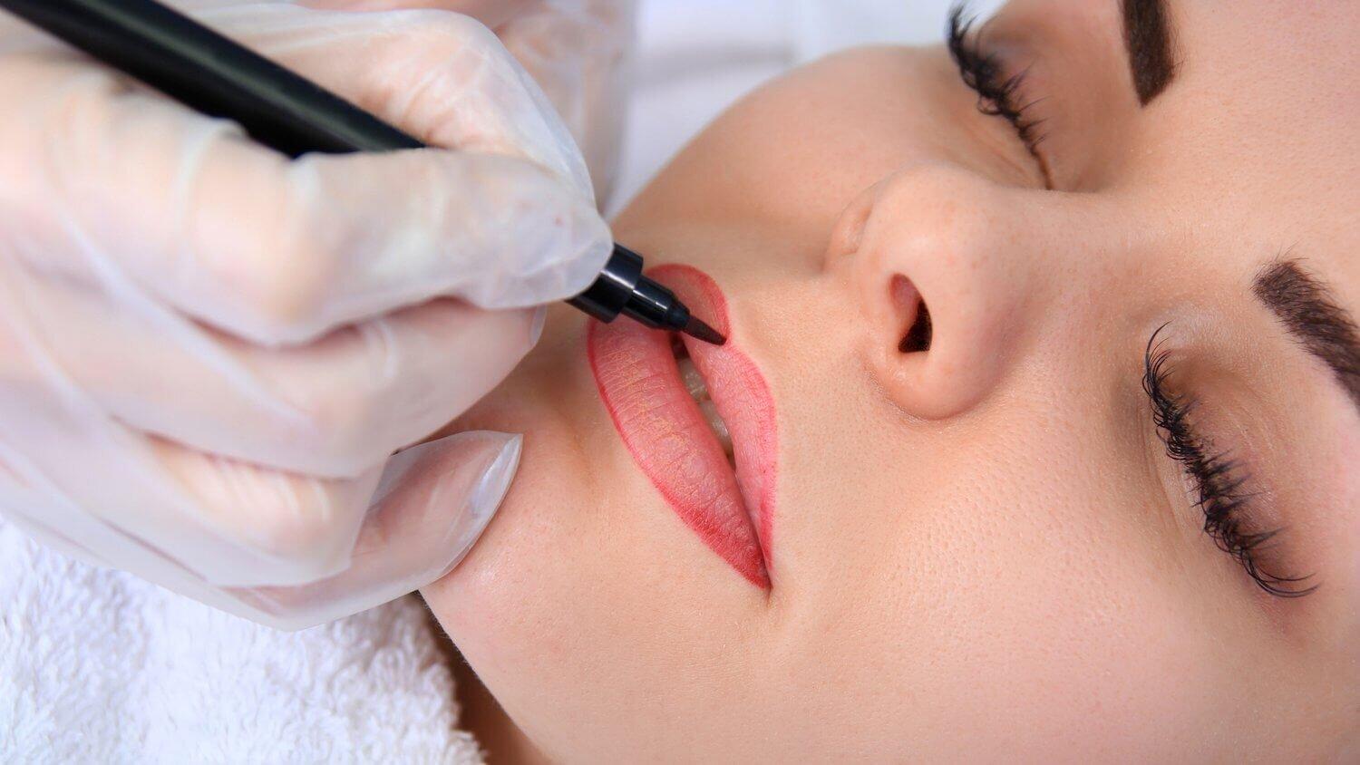 Das Bild zeigt eine Nahaufnahme einer Frau, die eine Permanent-Make-up-Behandlung für die Lippen erhält. Die Künstlerin, deren Hand mit einem Handschuh geschützt ist, trägt präzise rote Farbe auf die Lippenkontur der Kundin auf. Die Augen der Frau sind geschlossen, was darauf hindeutet, dass sie sich entspannt, während die Behandlung durchgeführt wird. Diese professionelle Permanent-Make-up-Behandlung wird von Dermacare Wien angeboten, wo höchste Präzision und individuelle Anpassung der Farbwahl für ein perfektes und langanhaltendes Ergebnis im Vordergrund stehen.