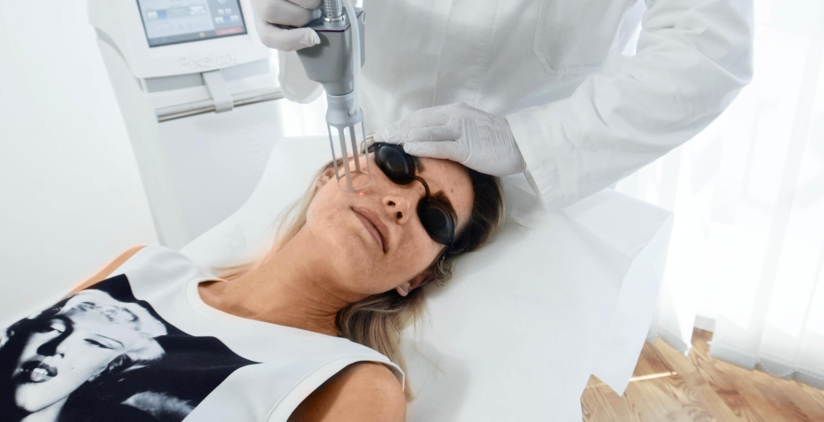 Auf dem Bild ist eine ästhetisch-medizinische Behandlung in einer professionellen Praxis zu sehen. Eine Patientin liegt entspannt auf einer Behandlungsliege, während ein medizinischer Spezialist mit Handschuhen ein modernes Lasersystem im Gesicht der Patientin anwendet. Die Patientin trägt eine Schutzbrille, um ihre Augen während der Lasertherapie zu schützen. Bei der Behandlung handelt es sich um eine schonende und präzise Laseranwendung, wie sie auch bei Dermacare Wien eingesetzt wird, um Pigmentflecken entfernen zu lassen und den Teint zu verfeinern. Der Hintergrund ist klinisch sauber und modern, was auf hohe medizinische Standards und professionelle Hautbehandlungen hinweist.