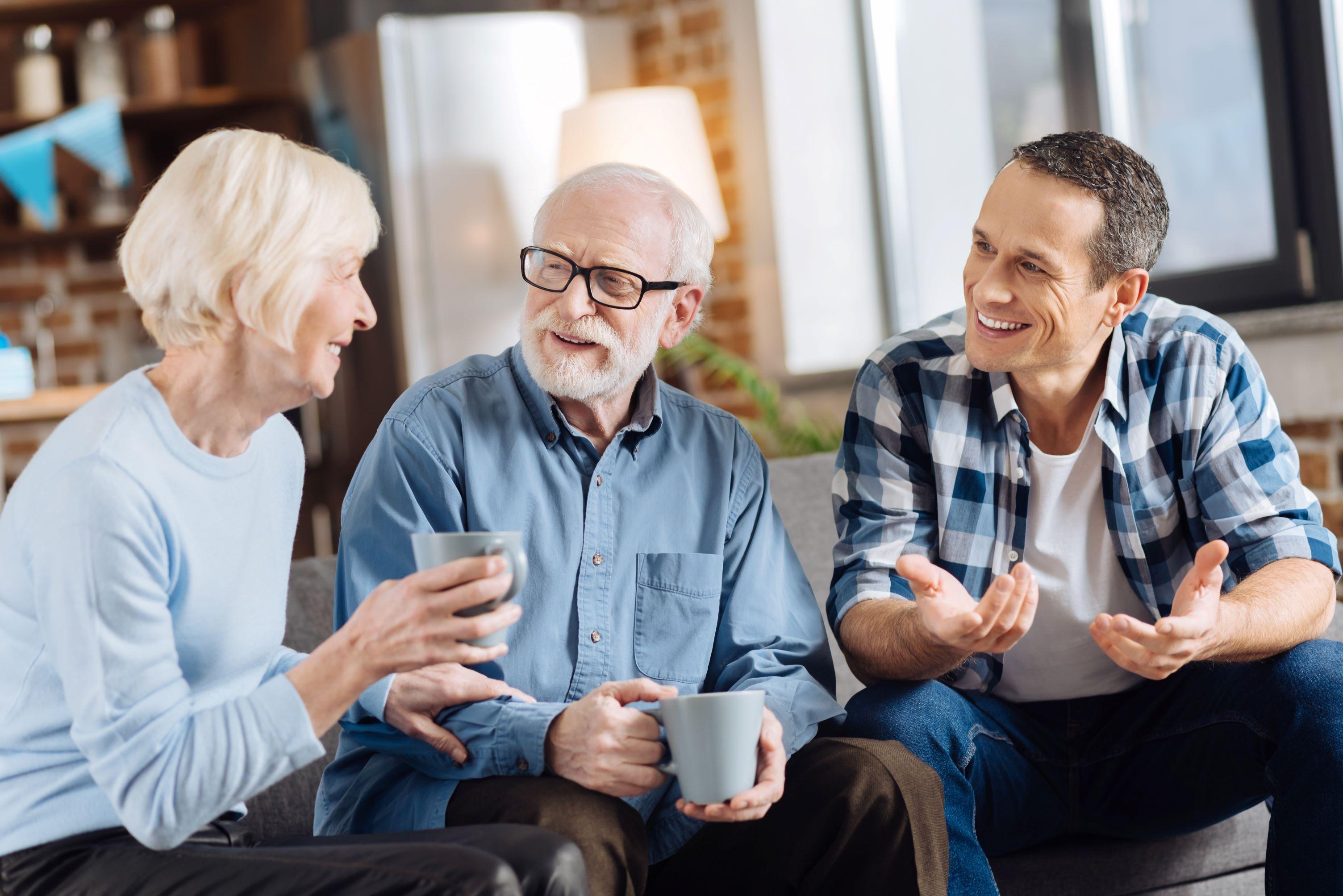 Cheerful senior couple sit with adult son drinking tea.