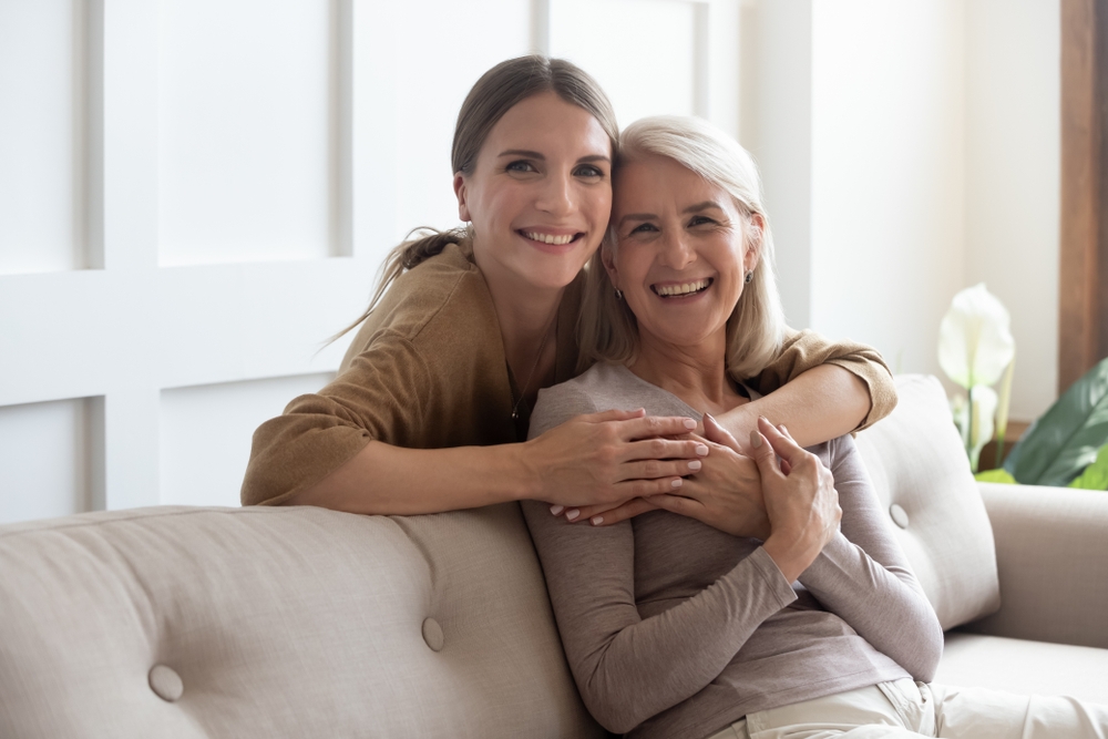 Loving adult daughter hugs her senior mother from behind while mom sits on couch.