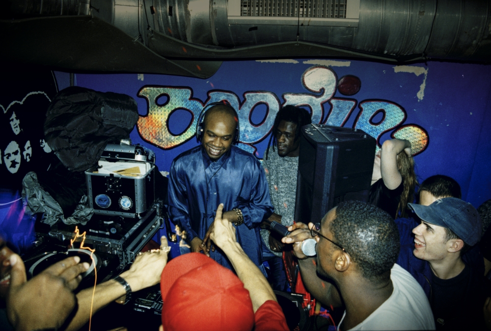 A DJ performing in a vibrant nightlife setting, engaged with a lively crowd, surrounded by turntables and audio equipment, against a colorful backdrop.