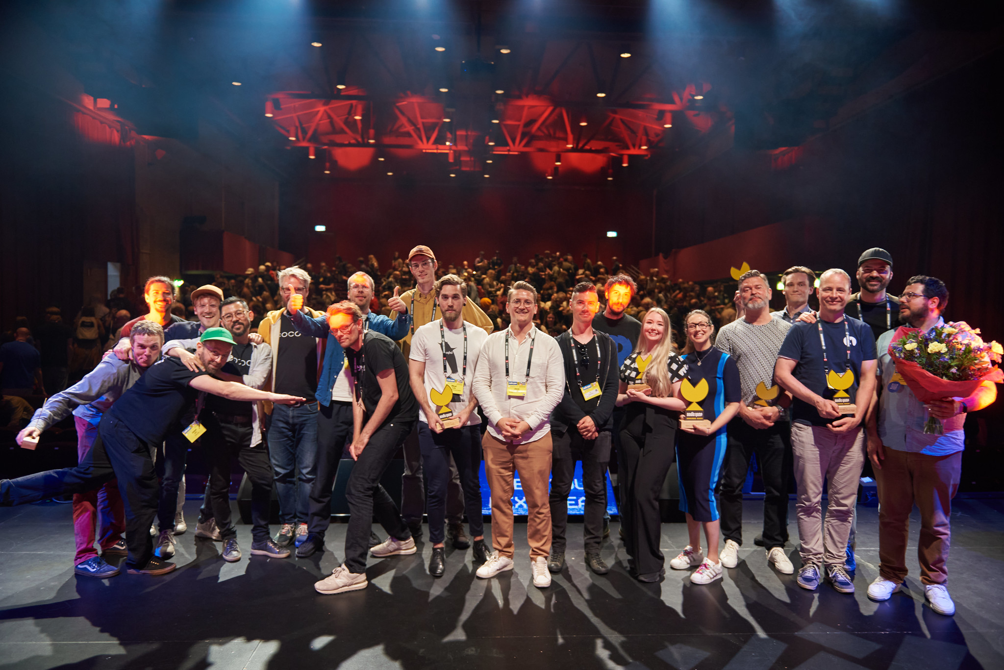 Foto. Gruppe på cirka 20 kvinner og menn står på en scene foran en fullsatt sal.