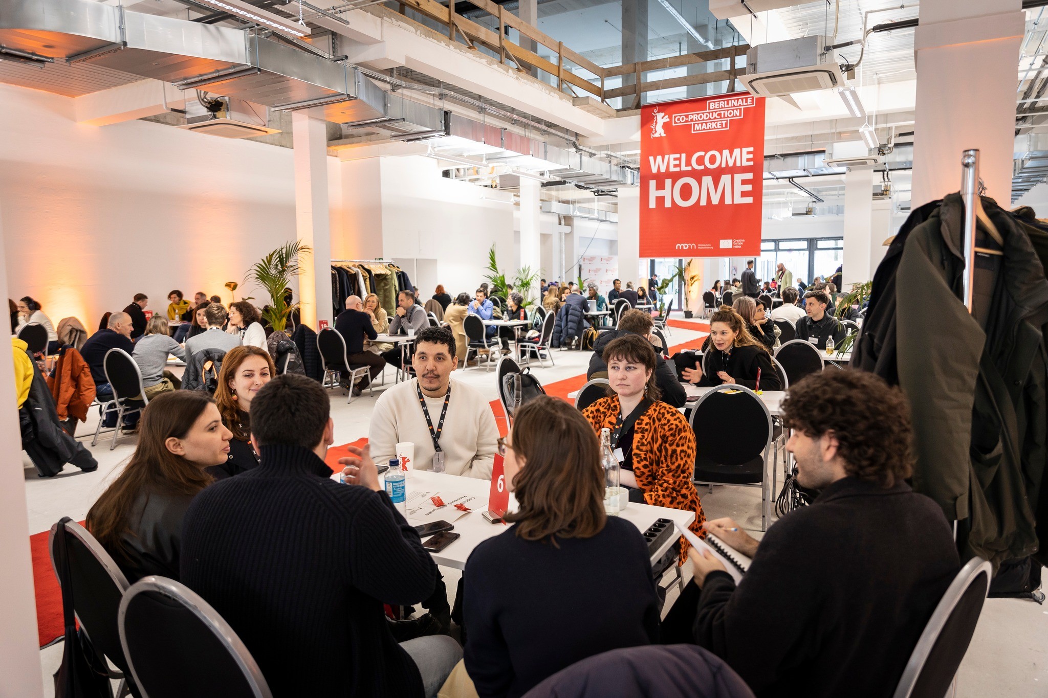 foto. bilde av en messehall med folk sittende rundt småbord under banner med påskriften Berlinale Coproduction Market