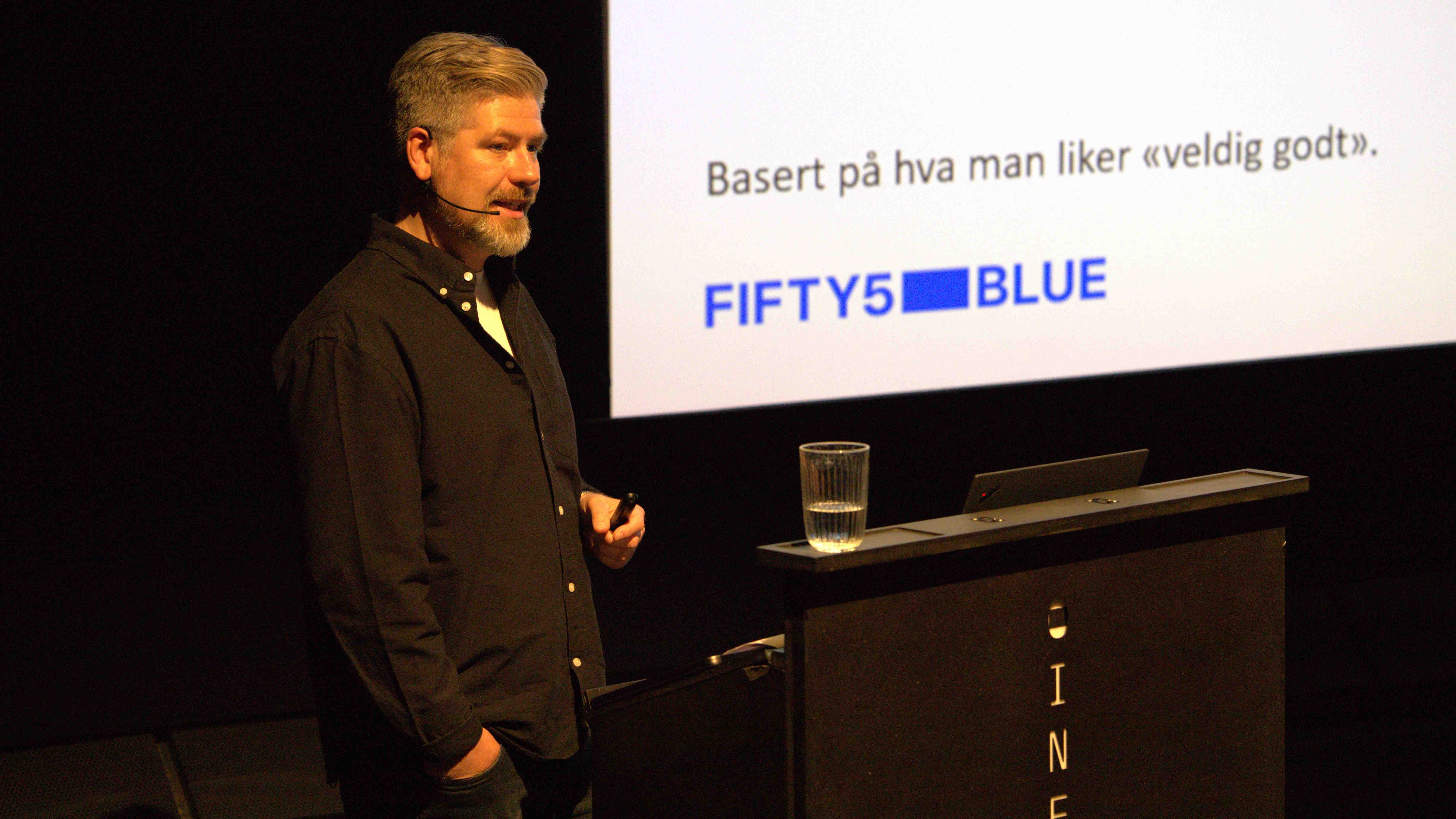 person med svart skjorte står ved Cinemaketets talerstol foran skjerm med påskriften Basert på hva man liker veldig godt, og logoen Fifty5 Blue