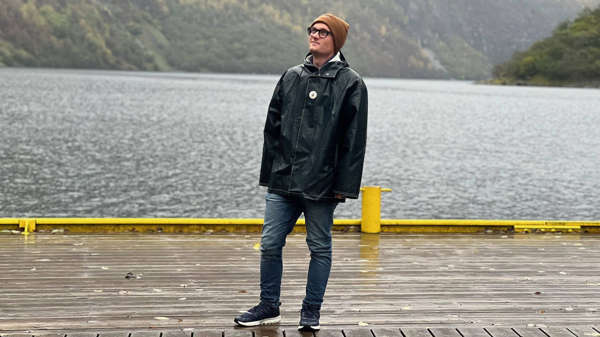 Foto. Mann står på en brygge med vann og fjell i bakgrunnen
