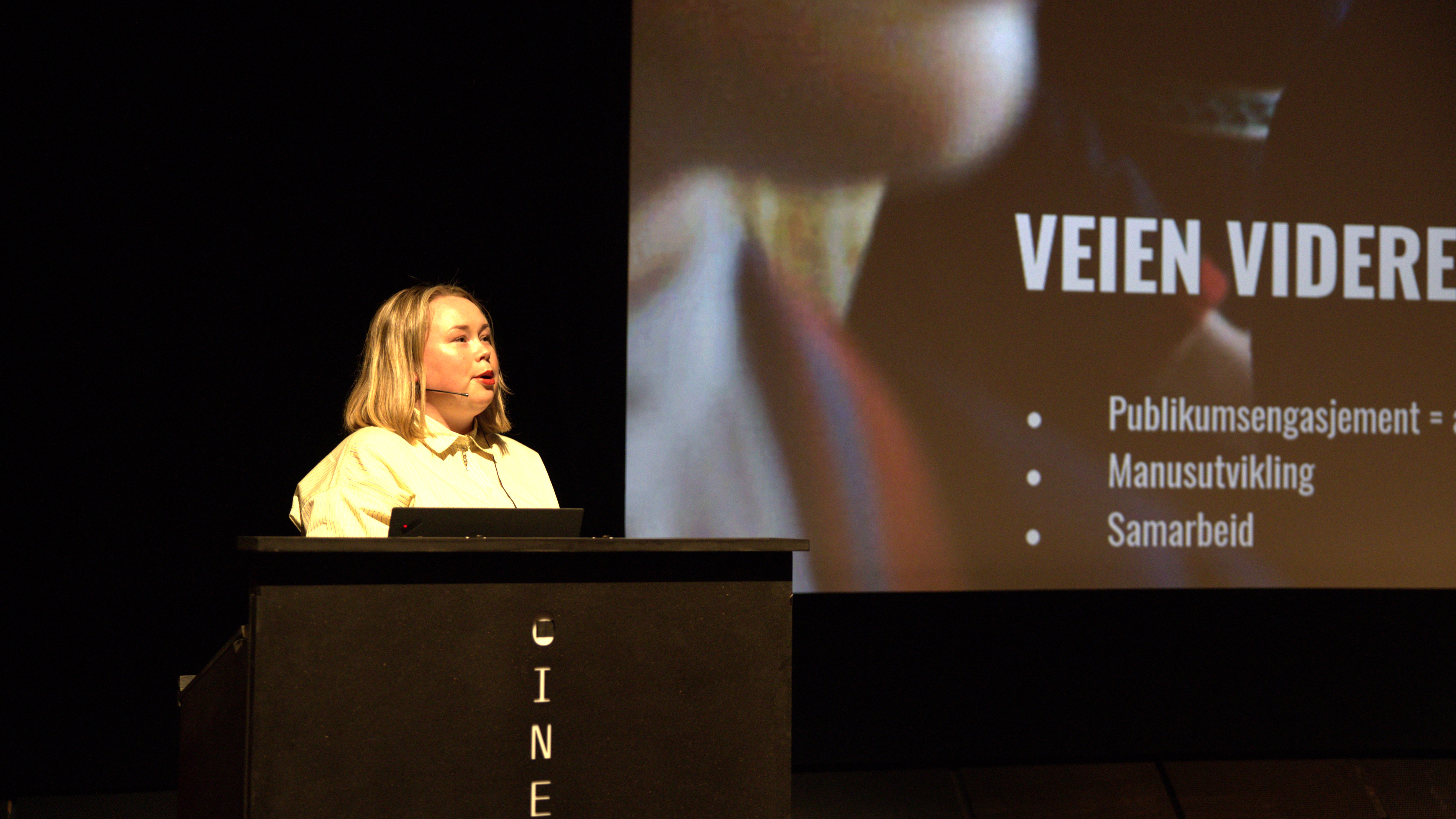 foto. ung kvinne står på Cinematekets talerstol med skjerm i bakgrunnen der det står Veien videre - publikumsengasjement manusutvikling